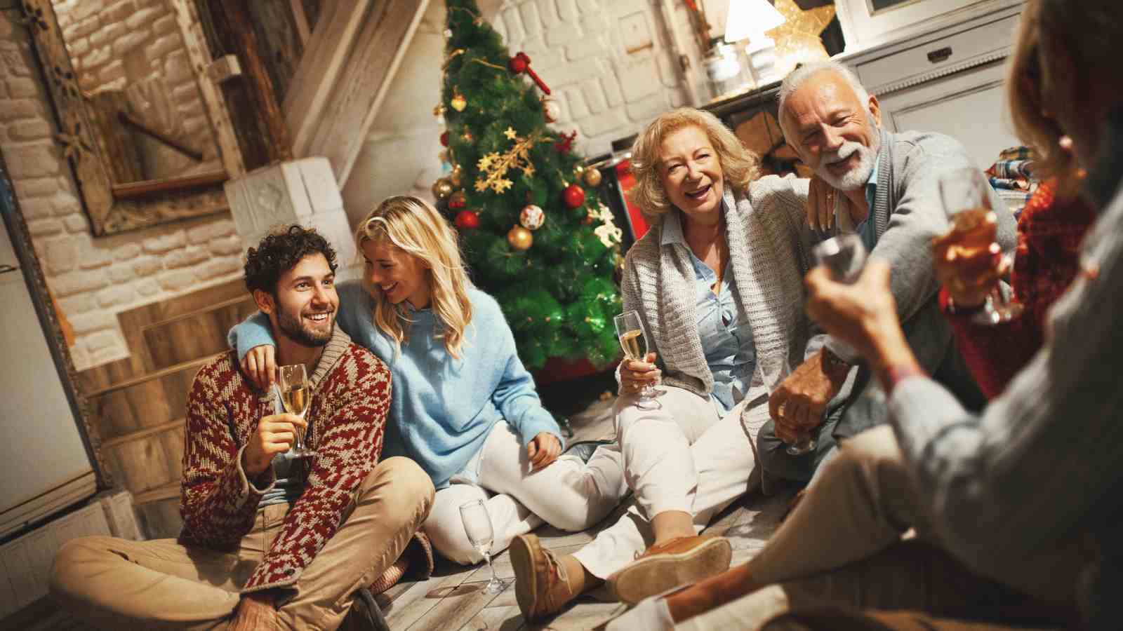 Five people sit on the floor near a decorated Christmas tree, smiling and holding drinks in a cozy indoor setting.
