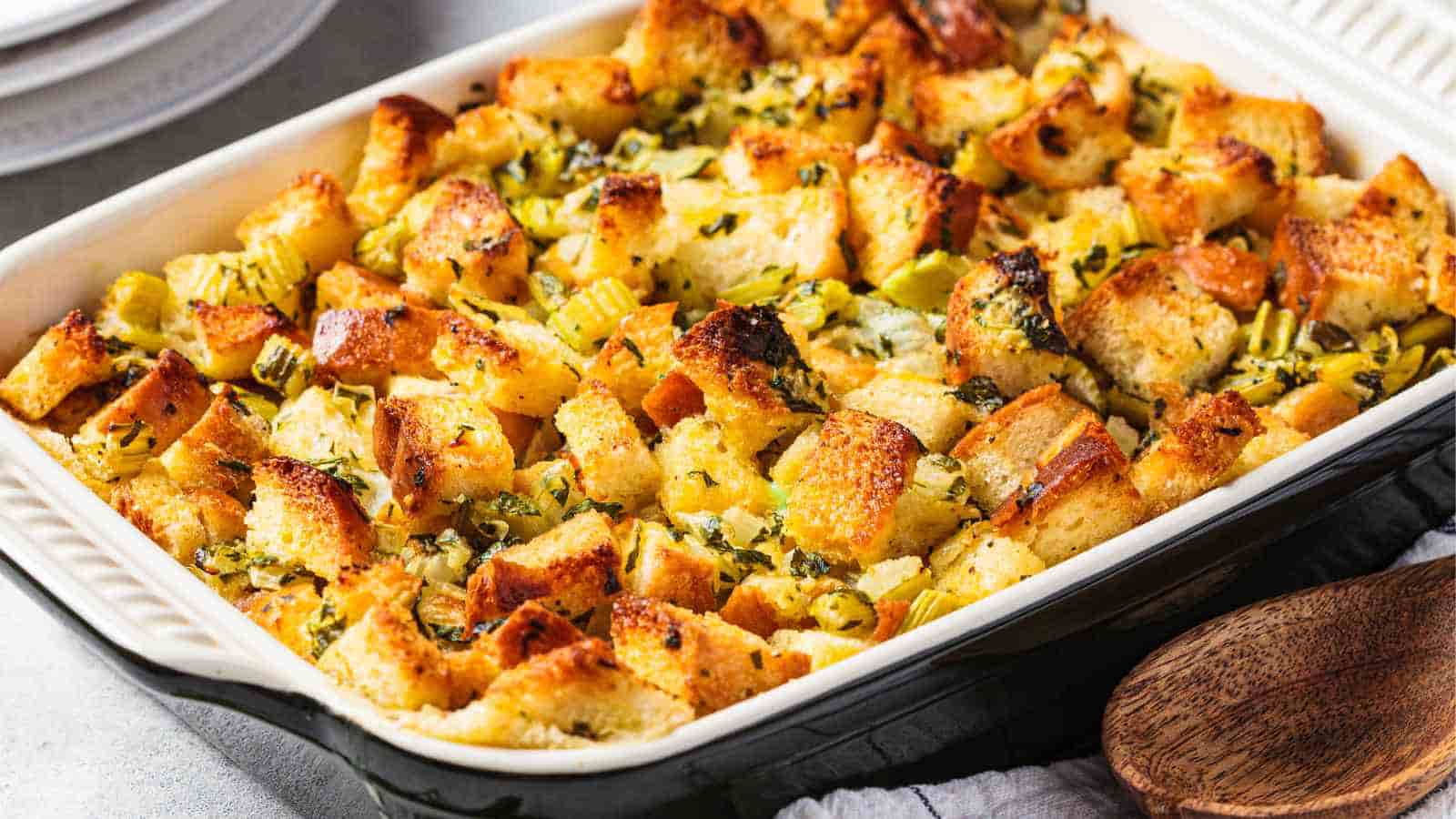A baking dish filled with golden-brown bread stuffing mixed with celery and herbs, with a wooden spoon beside it.