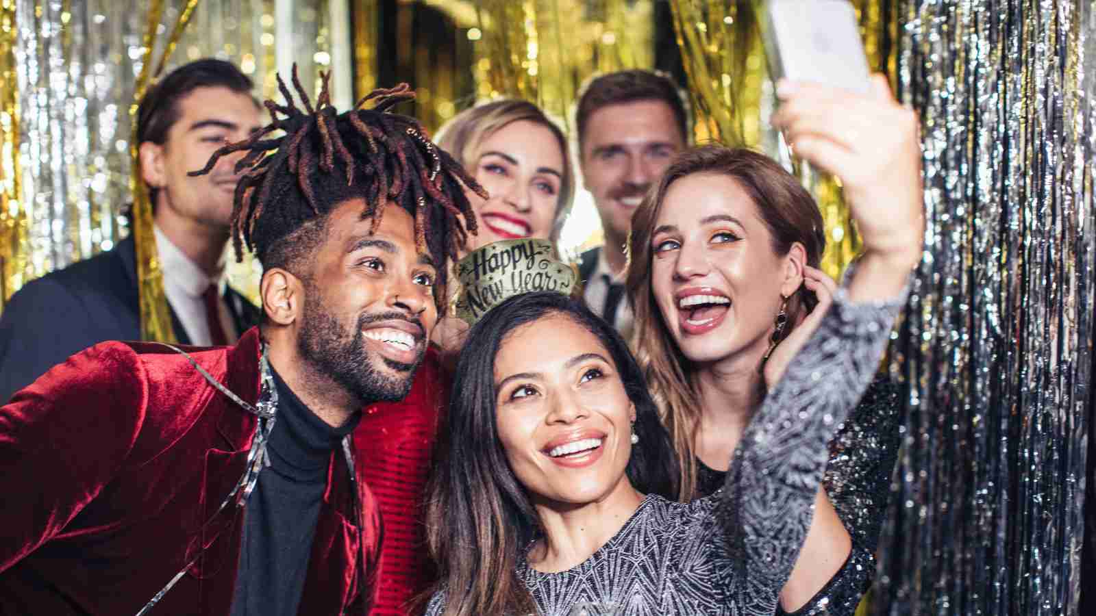 A group of six people smile and pose for a selfie at a festive party with gold and silver decorations in the background.