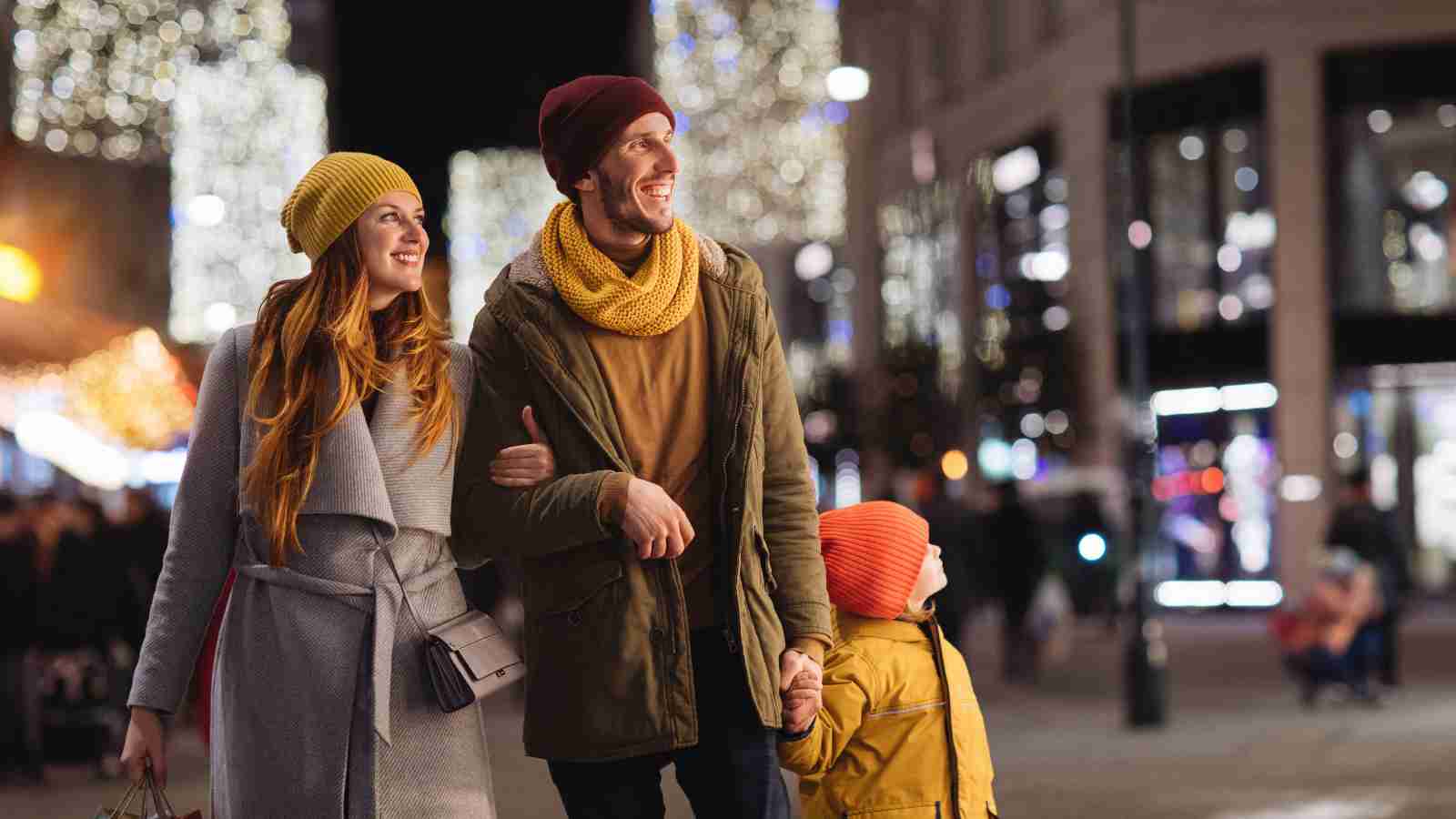 A family of three, dressed in winter clothing, walks together at night on a city street decorated with holiday lights.