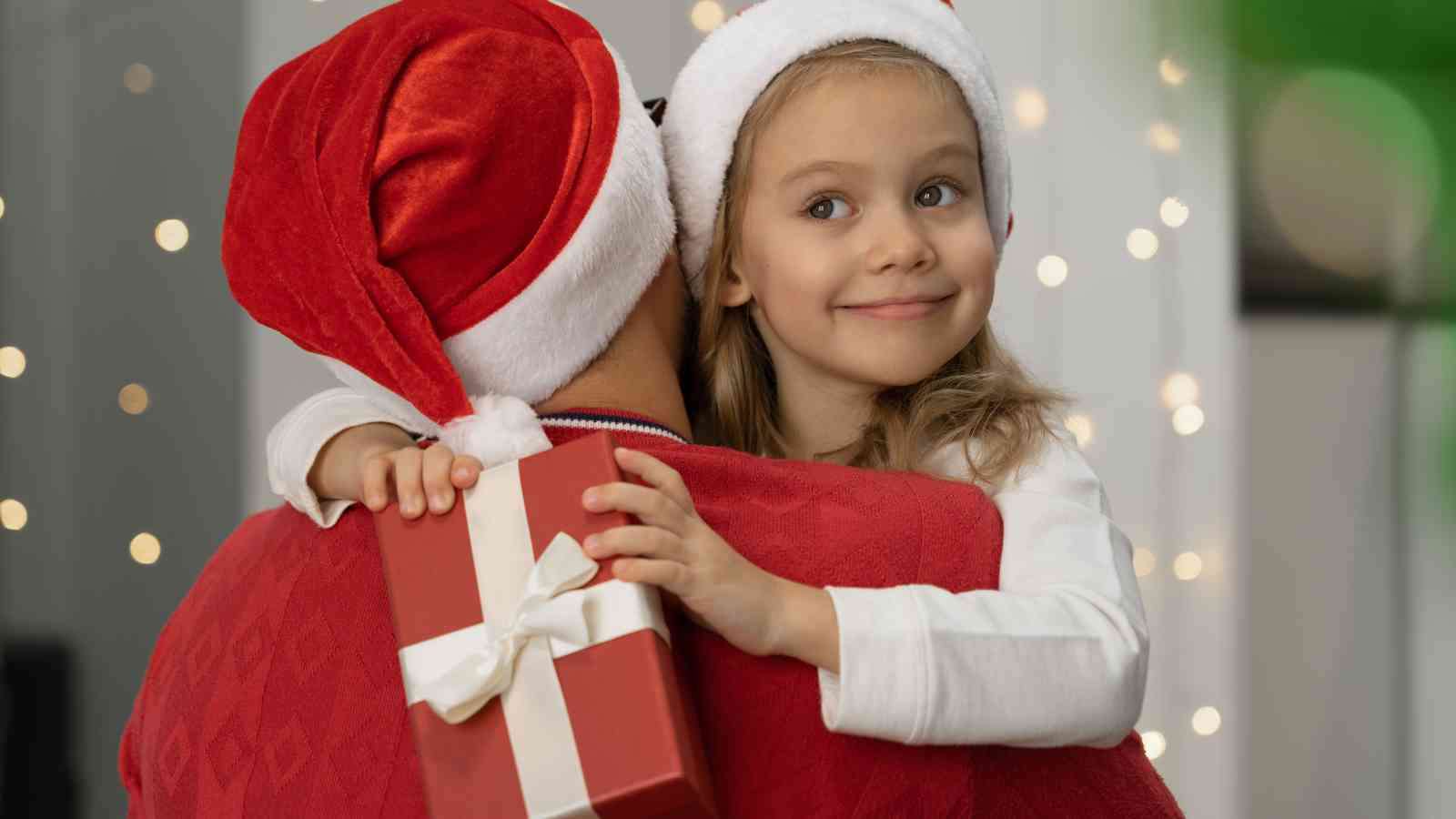 A young girl wearing a Santa hat hugs an adult in a Santa hat while holding a wrapped gift with a white ribbon.