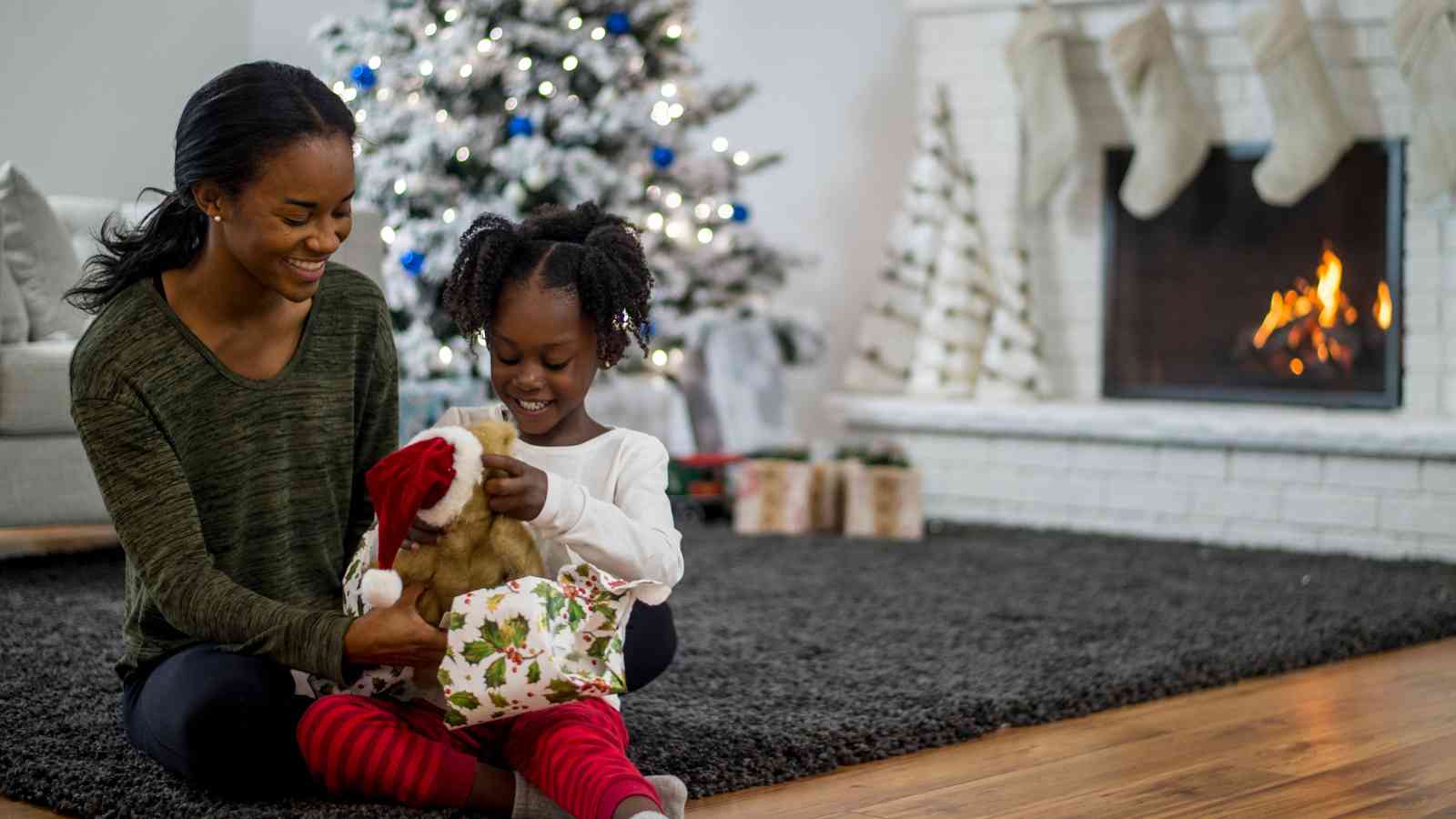A woman and child sit on a rug by a lit fireplace and Christmas tree, unwrapping a teddy bear in festive wrapping paper.