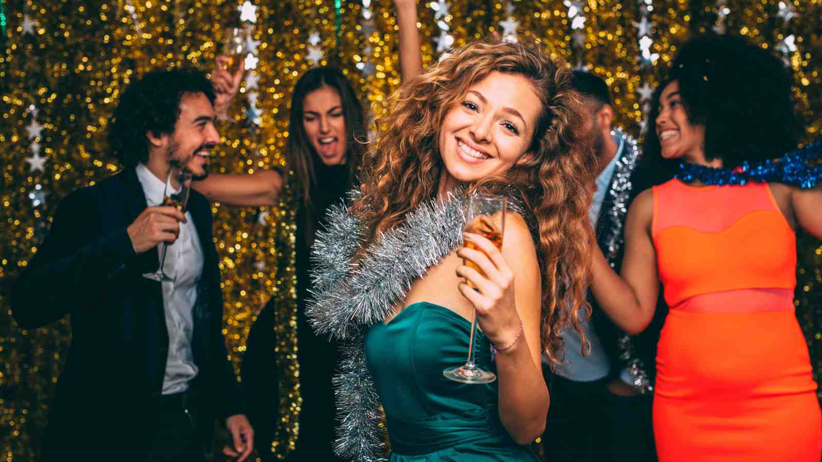 Five people dressed in party attire celebrate together, holding drinks and dancing in front of a gold tinsel backdrop. One woman in front is smiling at the camera.