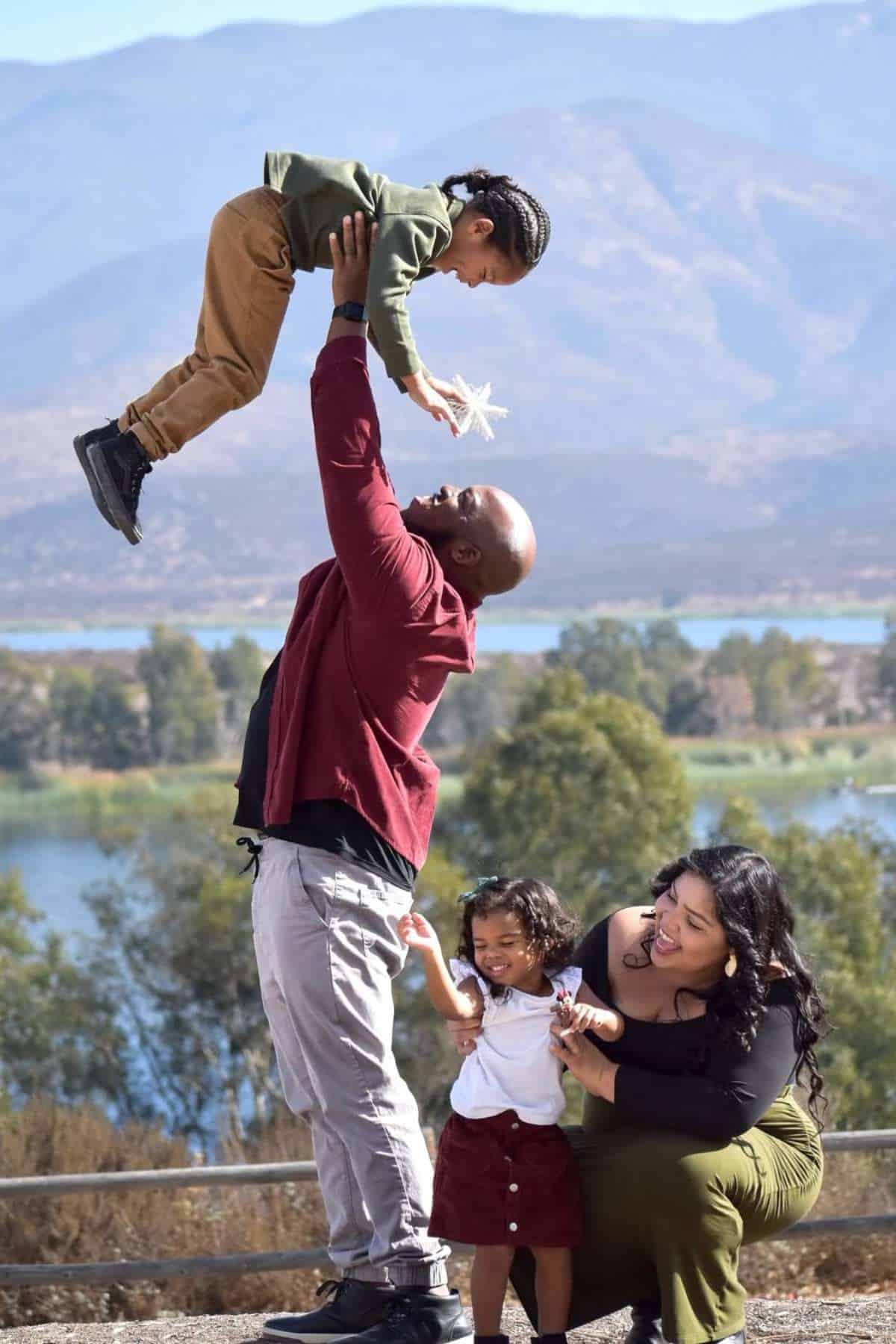 A man lifts a young child overhead while a woman kneels beside another child, outdoors with mountains and a lake in the background.