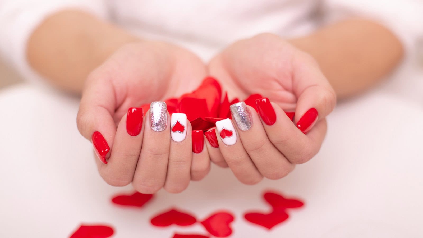 Hands with red, white, and silver manicured nails, some featuring heart designs, holding red heart-shaped confetti, with more confetti scattered on a white surface.