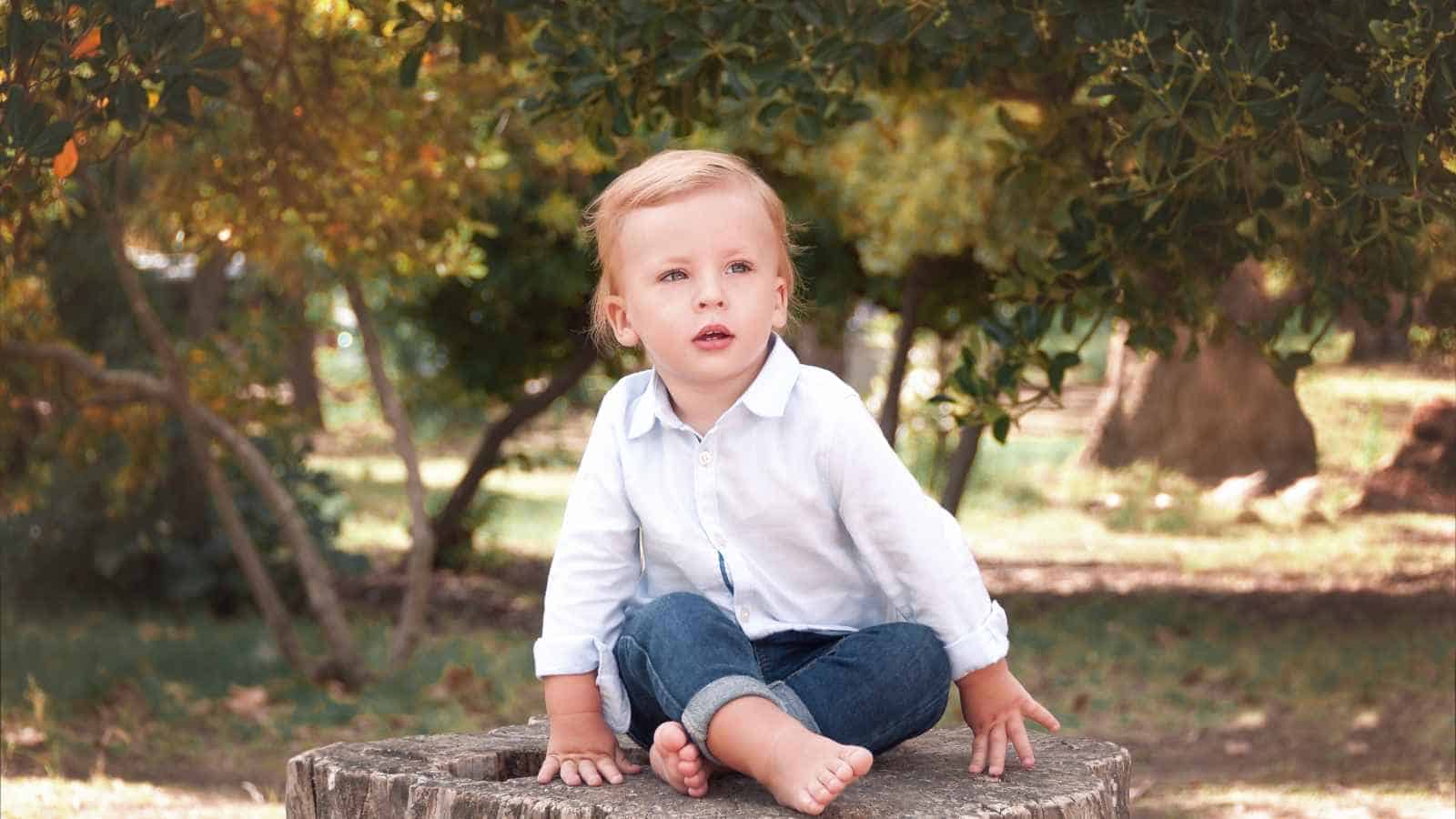 A young child with blond hair, wearing a white shirt and jeans, sits barefoot on a tree stump outdoors, surrounded by trees and greenery.