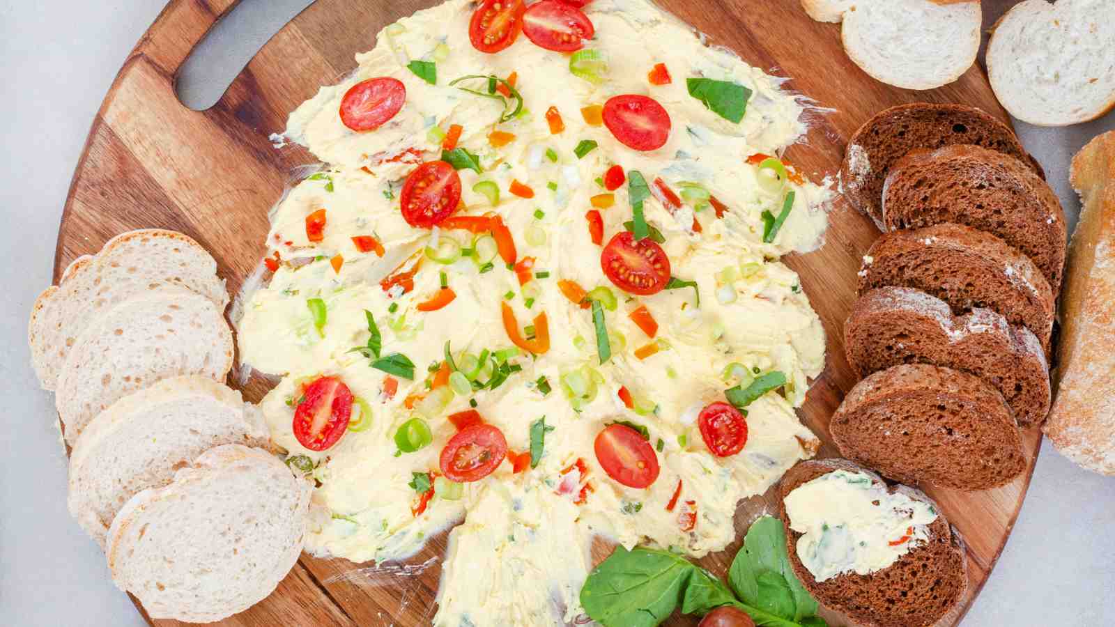 A wooden board with a Christmas tree-shaped spread topped with cherry tomatoes, chopped vegetables, and surrounded by slices of white and brown bread.