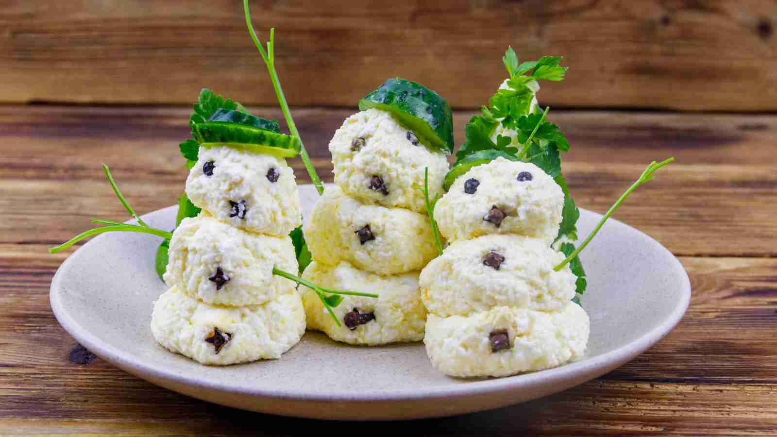 Three edible snowmen made from small stacks of cheese, decorated with herbs and peppercorns, are arranged on a plate against a wooden background.