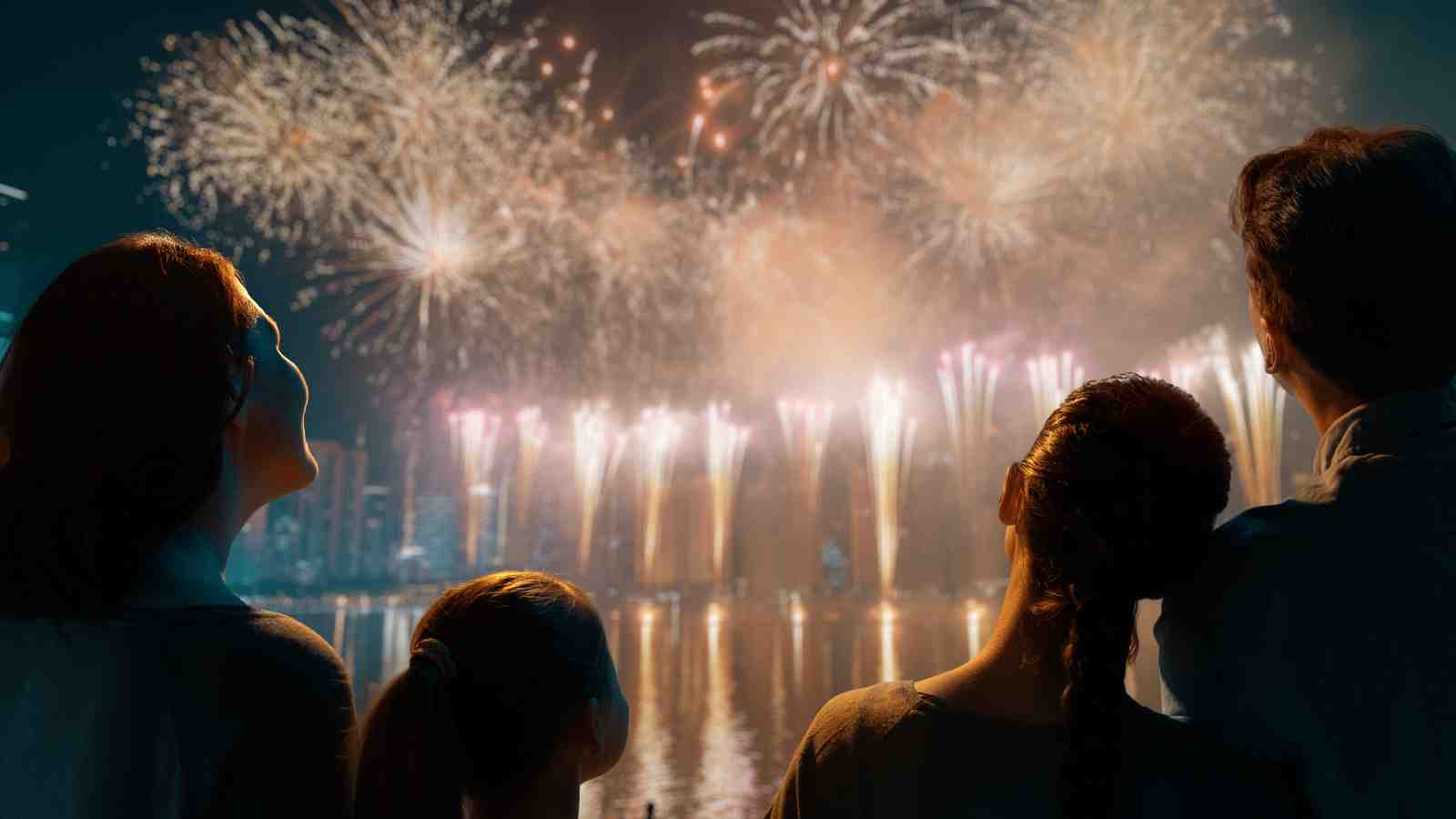 A family of four watches a colorful fireworks display over a city skyline at night, reflected in the water.