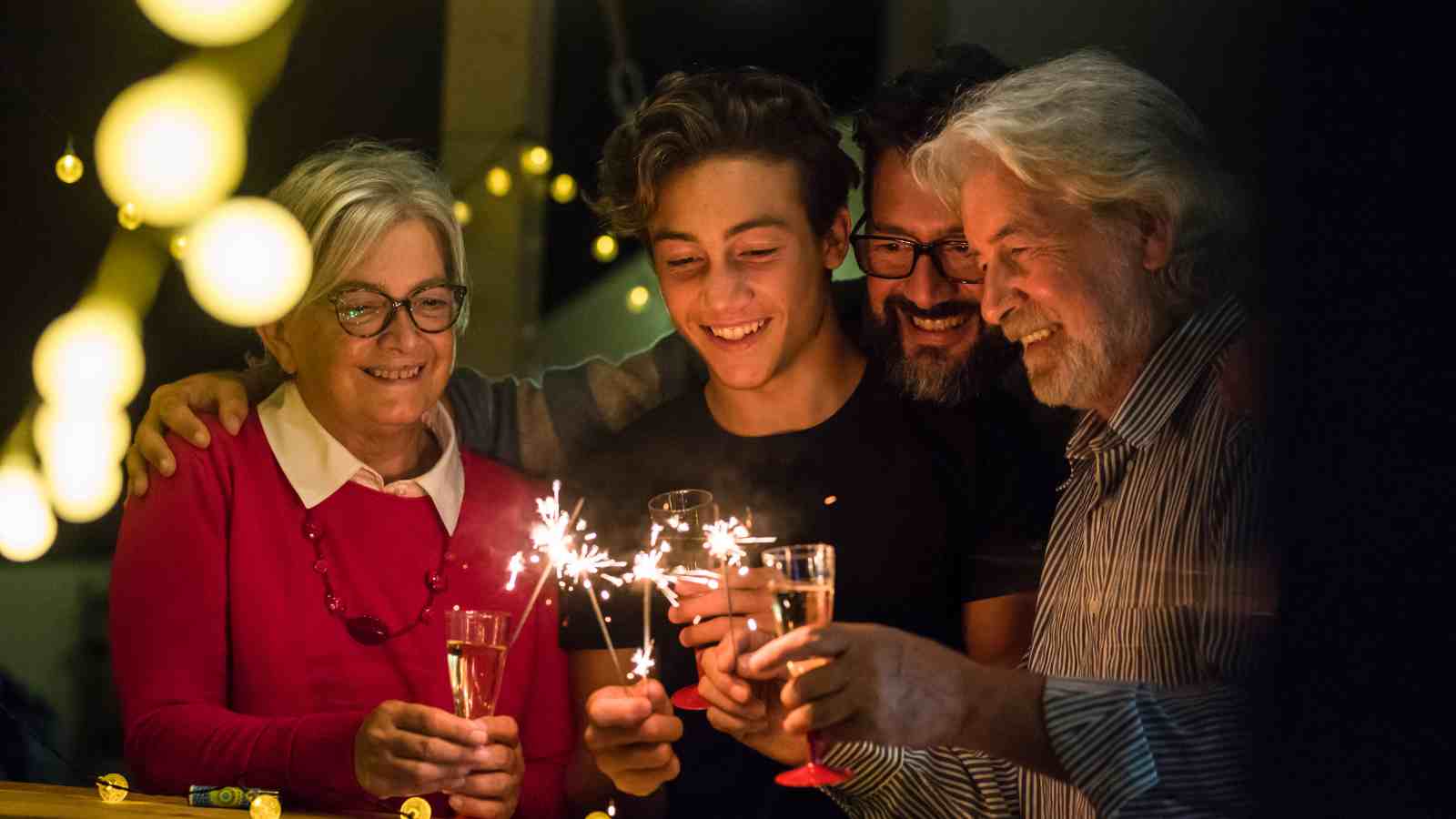 Four people stand close together, smiling and holding sparklers and glasses, celebrating in a warmly lit setting with string lights in the background.