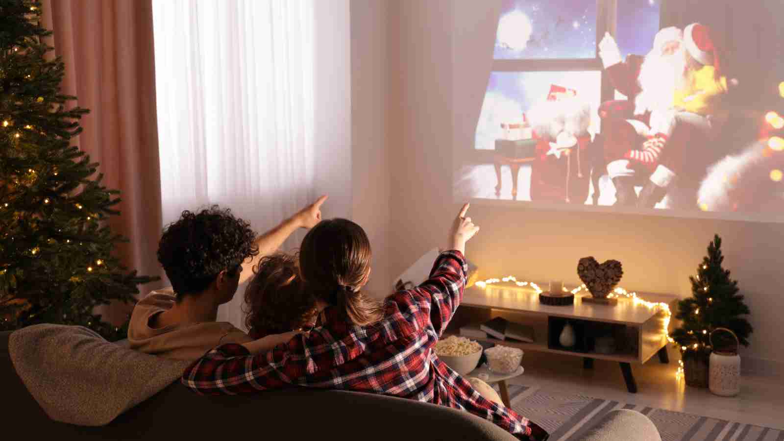 Two people sitting on a couch point at a Christmas-themed movie projected on a wall, surrounded by festive decorations including a Christmas tree and string lights.
