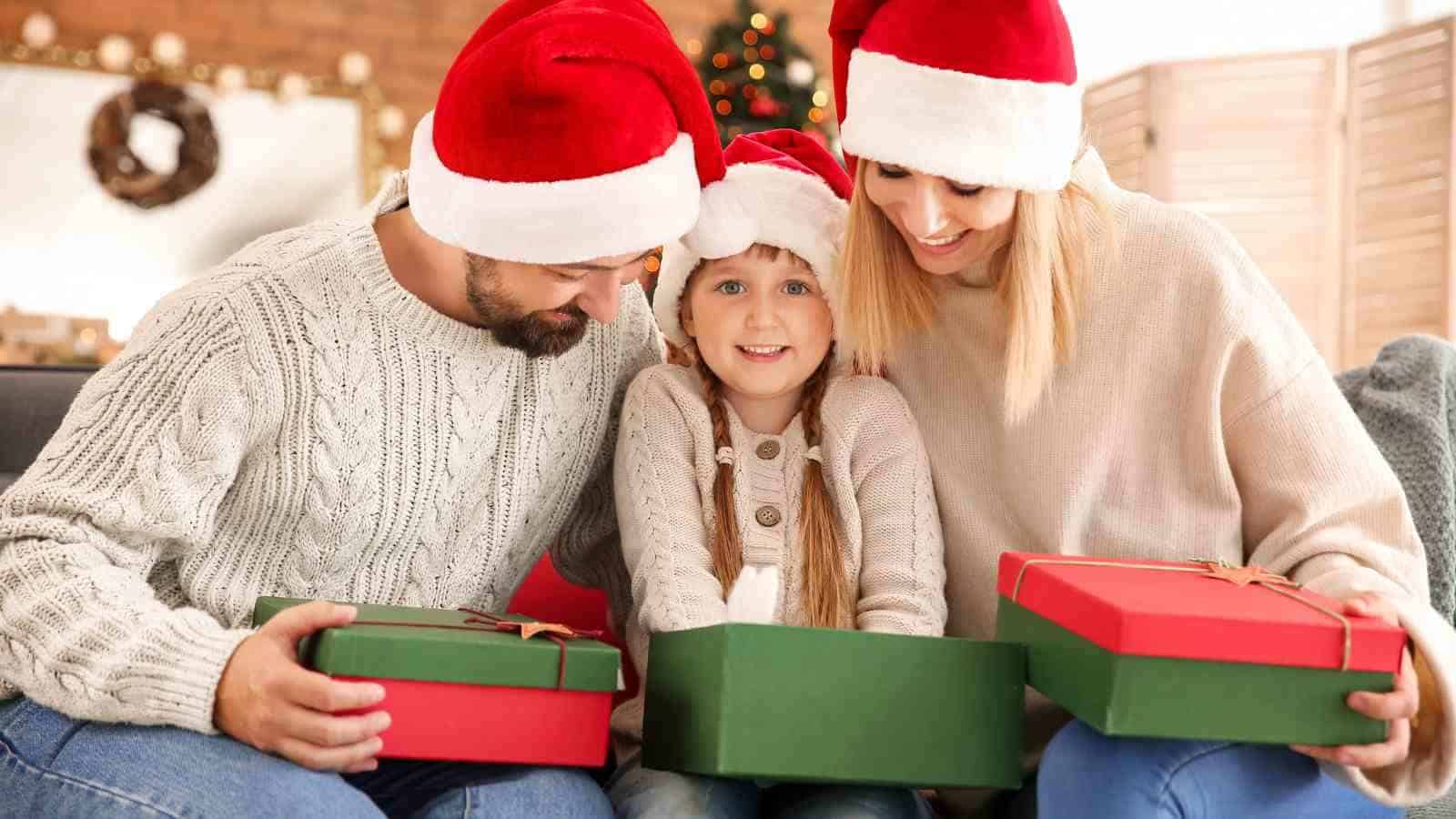 A family of three wearing Santa hats sits together on a couch, smiling as a child in the center opens a green gift box, with another gift box nearby.
