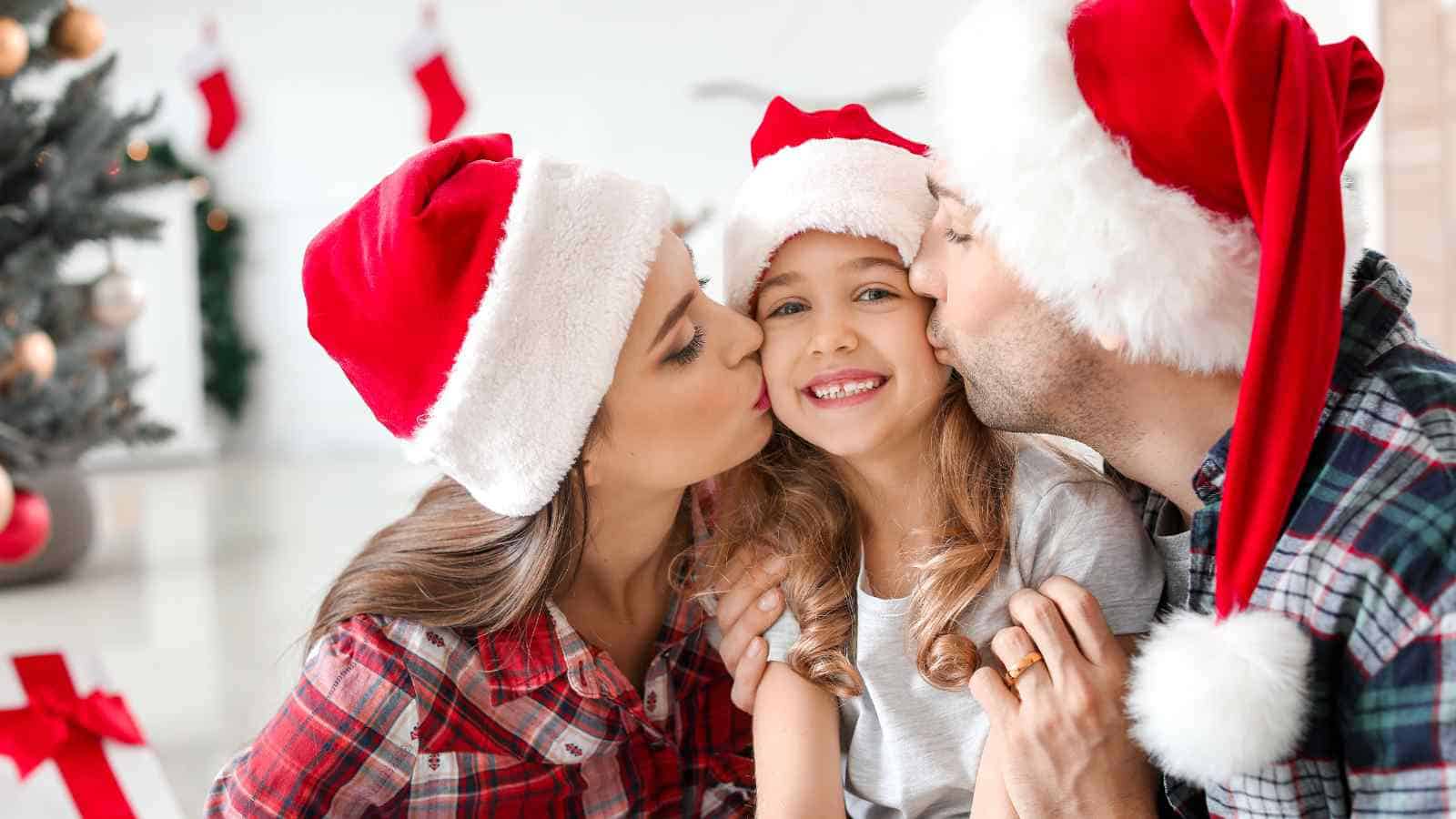 A smiling child wearing a Santa hat is kissed on the cheeks by two adults, also in Santa hats, in a festive room with a Christmas tree, stockings, and presents.