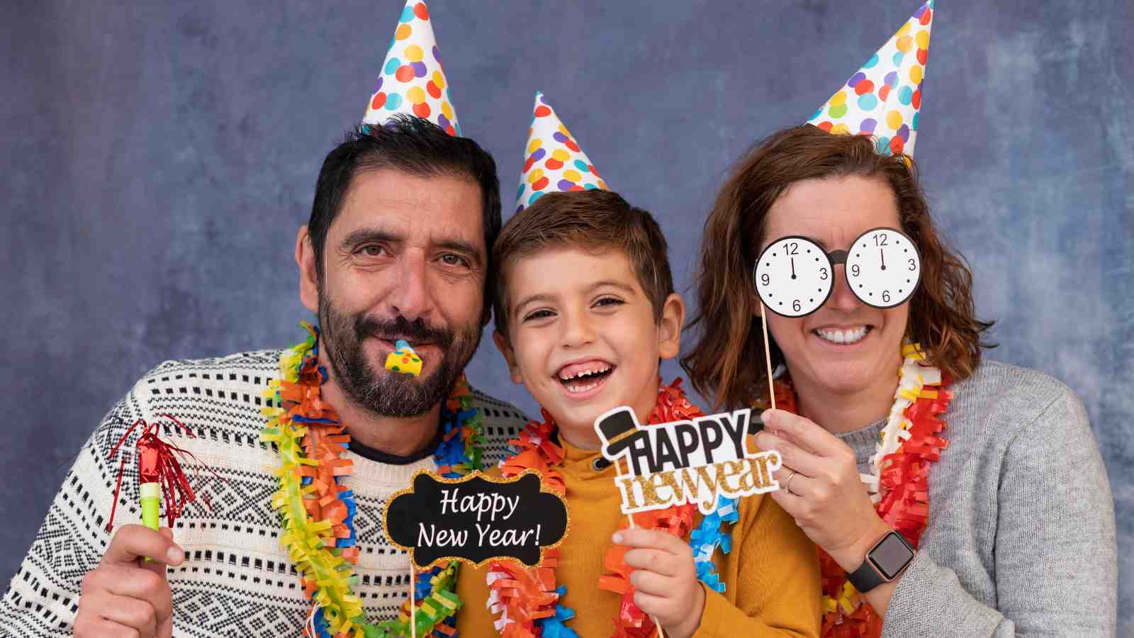 Three people wearing party hats and festive accessories smile at the camera, holding "Happy New Year" signs and celebrating together.
