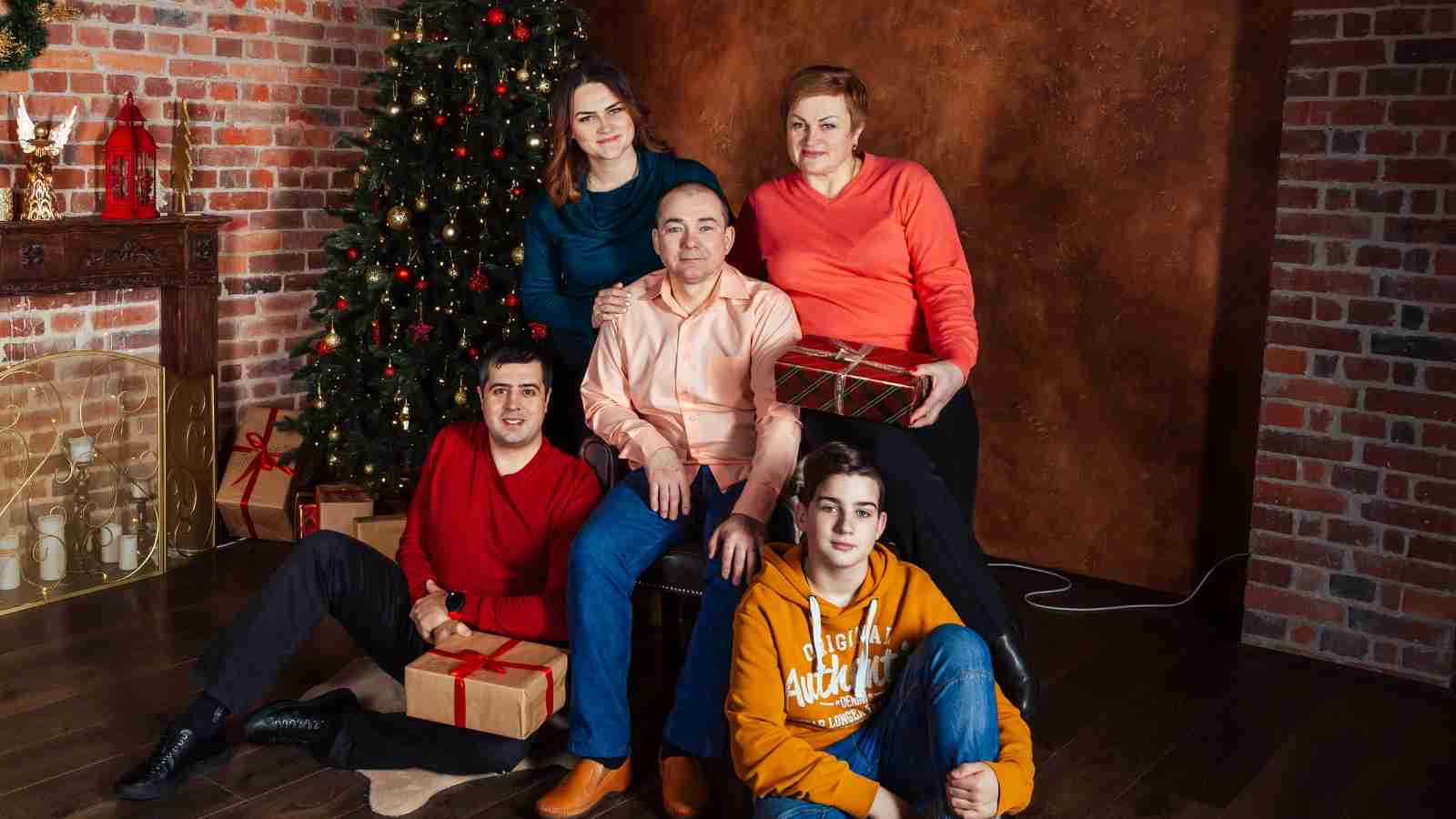 Five people pose in front of a decorated Christmas tree and wrapped gifts, sitting and standing together indoors with a brick wall background.