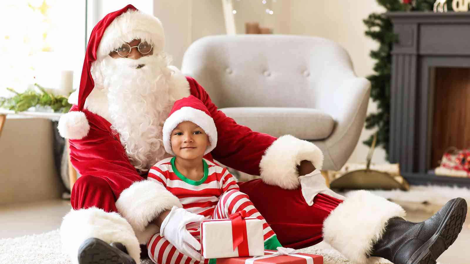 An adult dressed as Santa Claus sits on the floor with a young child in striped pajamas and Santa hat, surrounded by wrapped gifts in a living room decorated for Christmas.