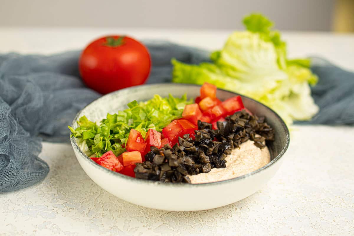 A bowl containing chopped lettuce, diced tomatoes, black olives, and a creamy dip sits on a light surface, with a whole tomato and lettuce in the background.
