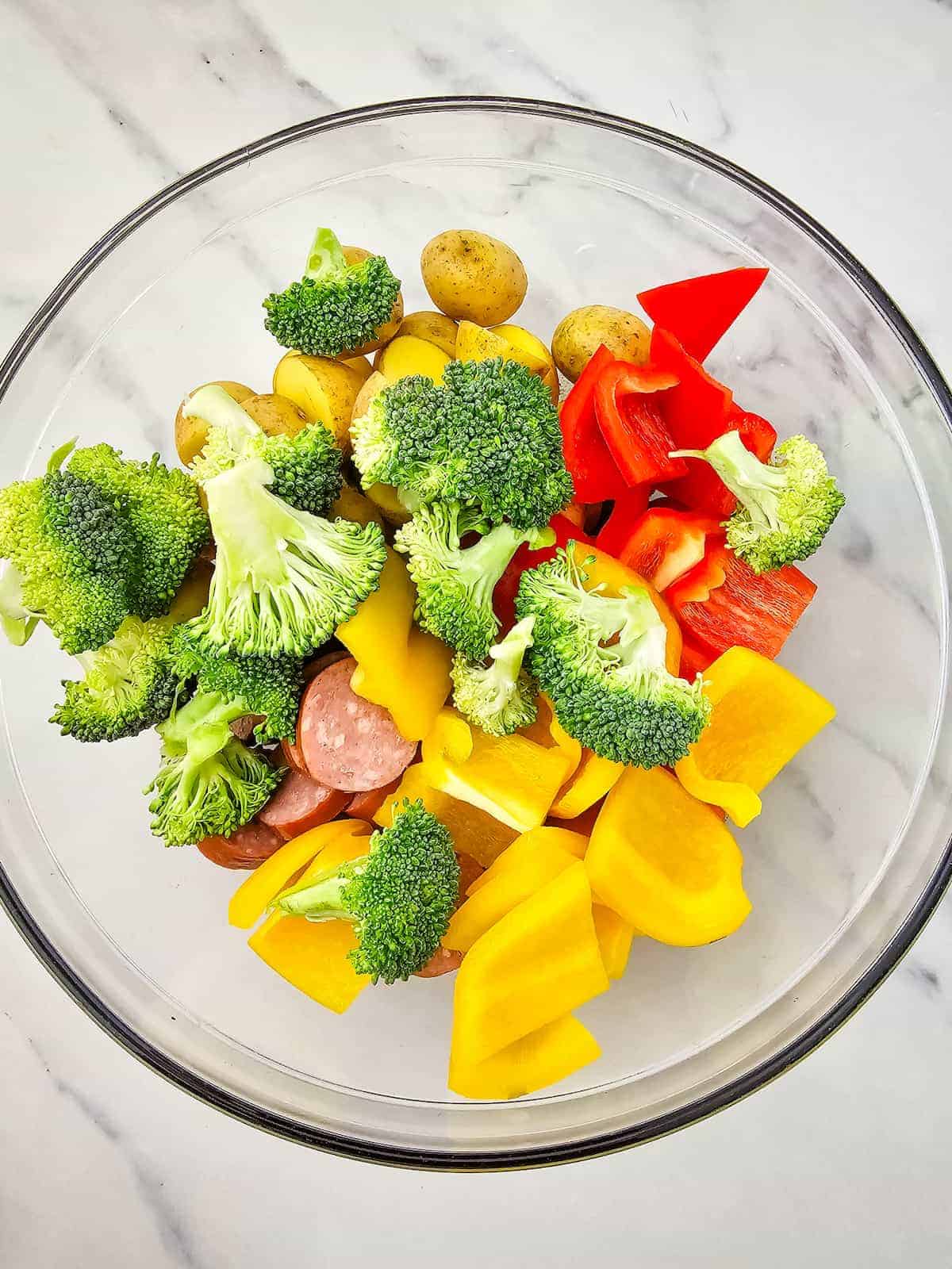 A glass bowl containing broccoli florets, red and yellow bell pepper chunks, sliced sausage, and small potatoes on a white marble surface.