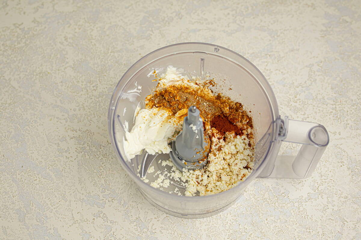 A food processor bowl containing cream cheese and spices ready to be blended.