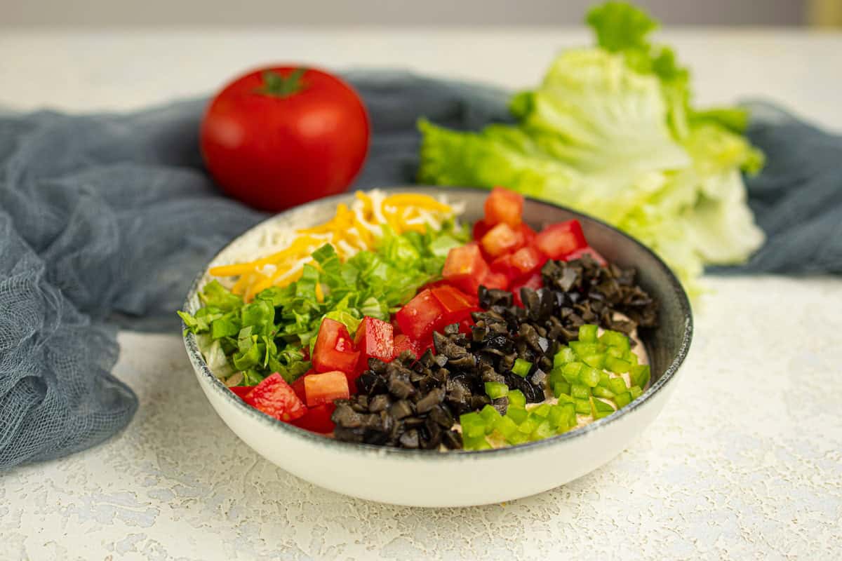 A bowl of dip with chopped lettuce, tomatoes, olives, green peppers, and shredded cheese, with a whole tomato and lettuce in the background.