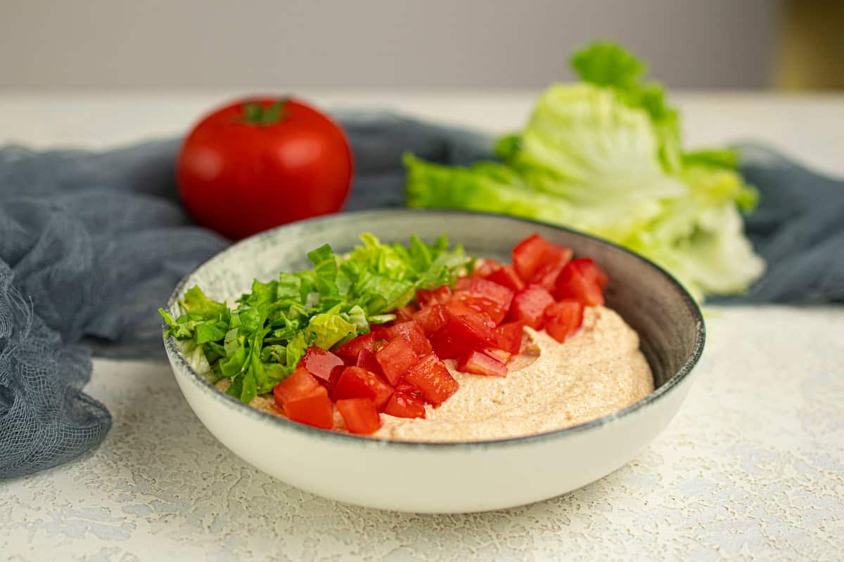 A bowl topped with chopped lettuce and diced tomatoes, with a whole tomato and lettuce leaves in the background.