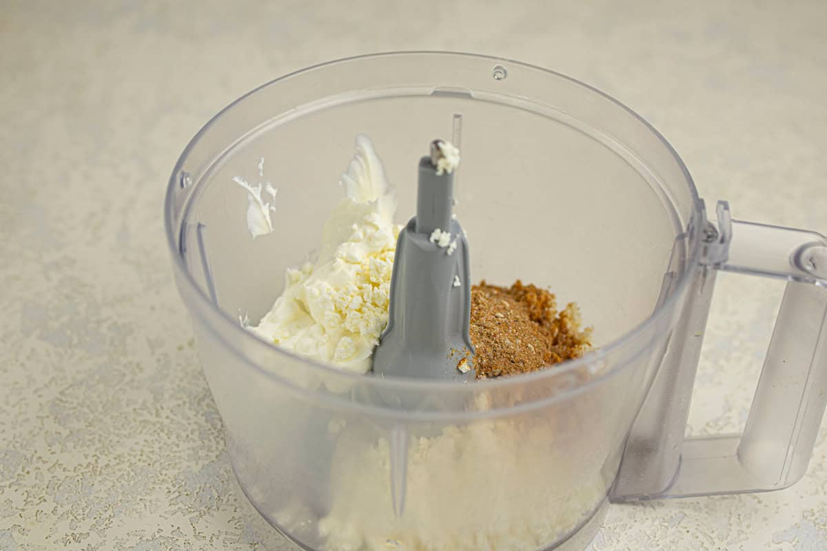 A food processor bowl containing cream cheese, grated cheese, and a taco seasoning mixture, ready to be blended.