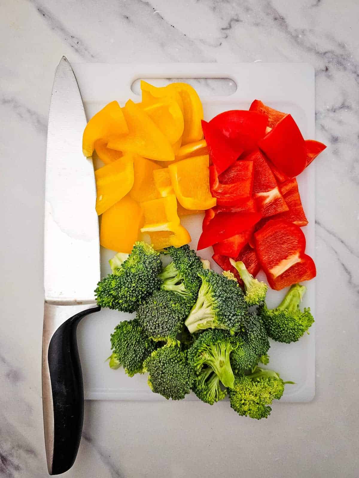 Chopped yellow bell pepper, red bell pepper, and broccoli florets are arranged on a white cutting board next to a kitchen knife.