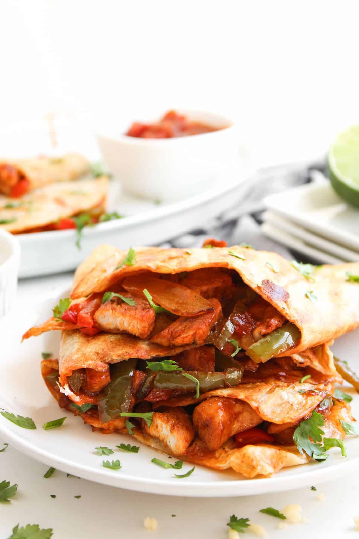 Two folded tortillas filled with seasoned chicken, bell peppers, and onions are served on a white plate, garnished with fresh cilantro. A bowl of salsa is in the background.