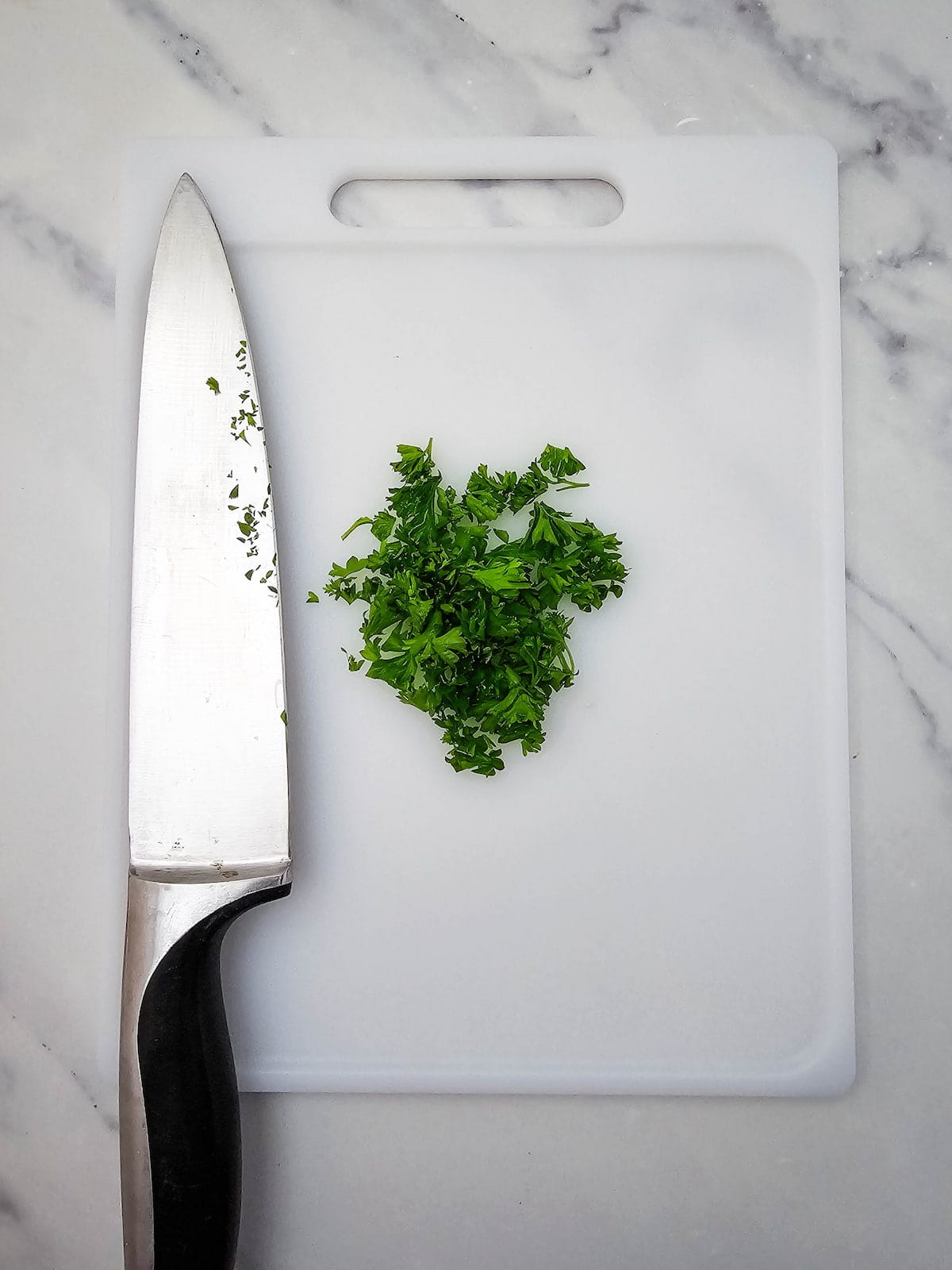 A chef’s knife and a small pile of chopped fresh parsley sit on a white cutting board atop a marble surface.