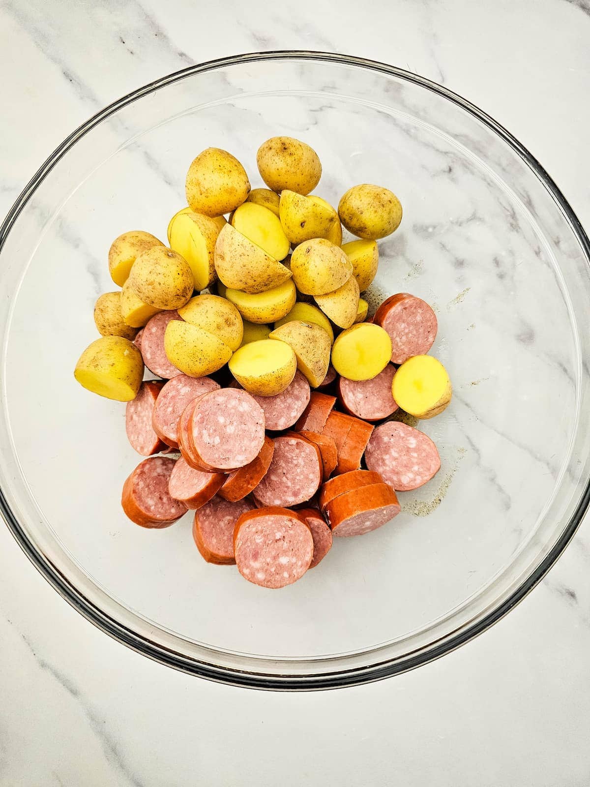 A glass bowl containing halved baby potatoes and sliced sausage on a white marble surface.