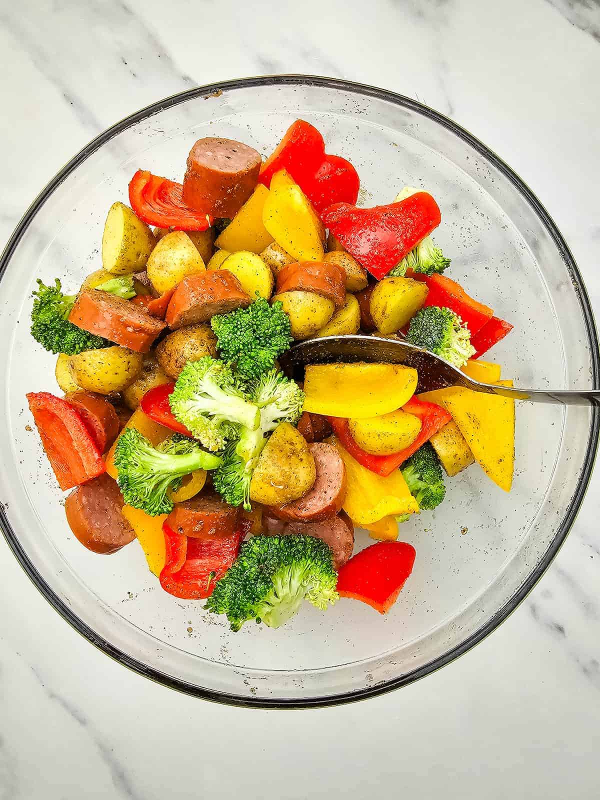 A glass bowl filled with sliced sausage, broccoli florets, bell peppers, and baby potatoes, with a metal spoon resting inside.