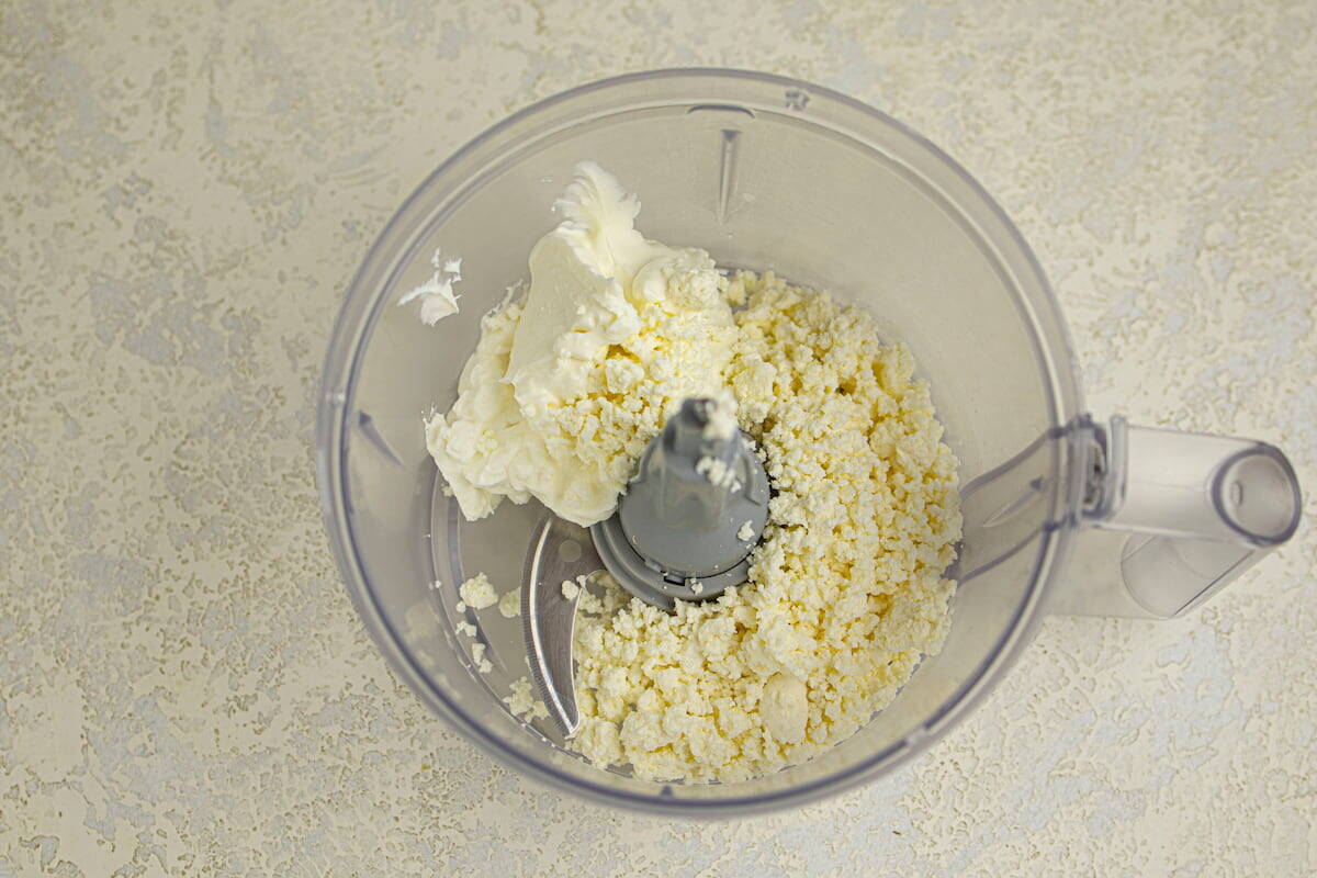 Crumbled cheese and a dollop of cream cheese in a food processor bowl on a light textured surface.