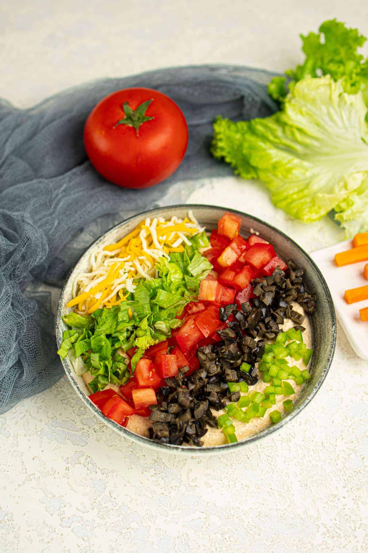 A bowl filled with layered dip topped with shredded cheese, chopped lettuce, tomatoes, black olives, and green bell peppers. A whole tomato and lettuce are in the background.