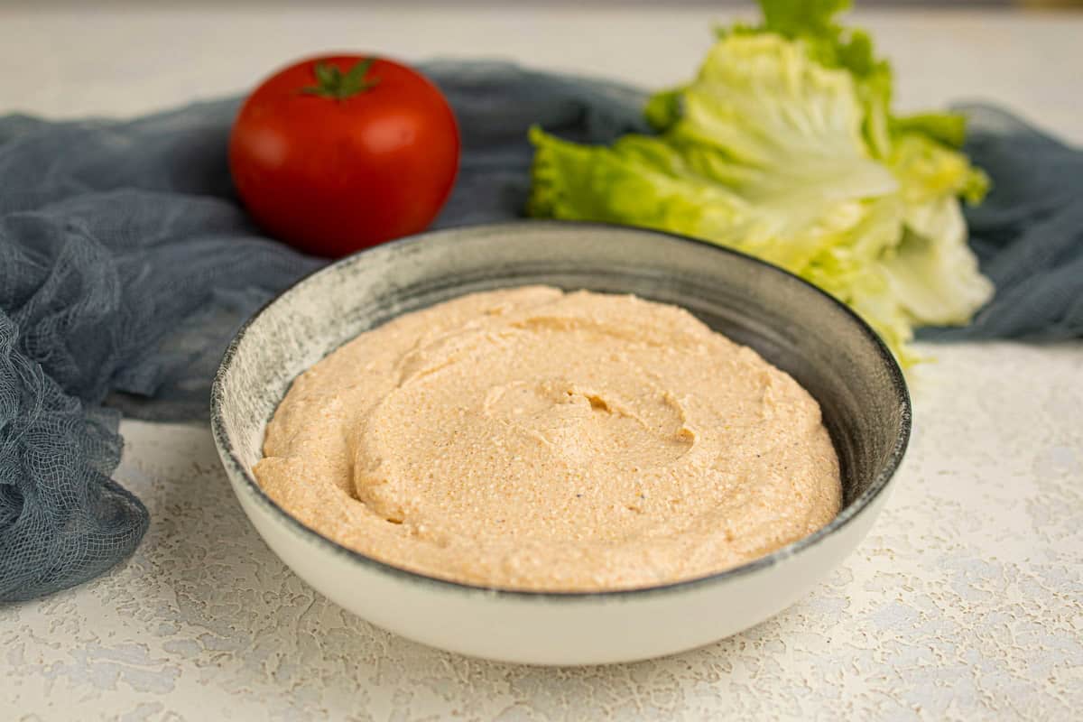A bowl of creamy dip sits on a light-colored surface, with a tomato and leafy lettuce in the background.