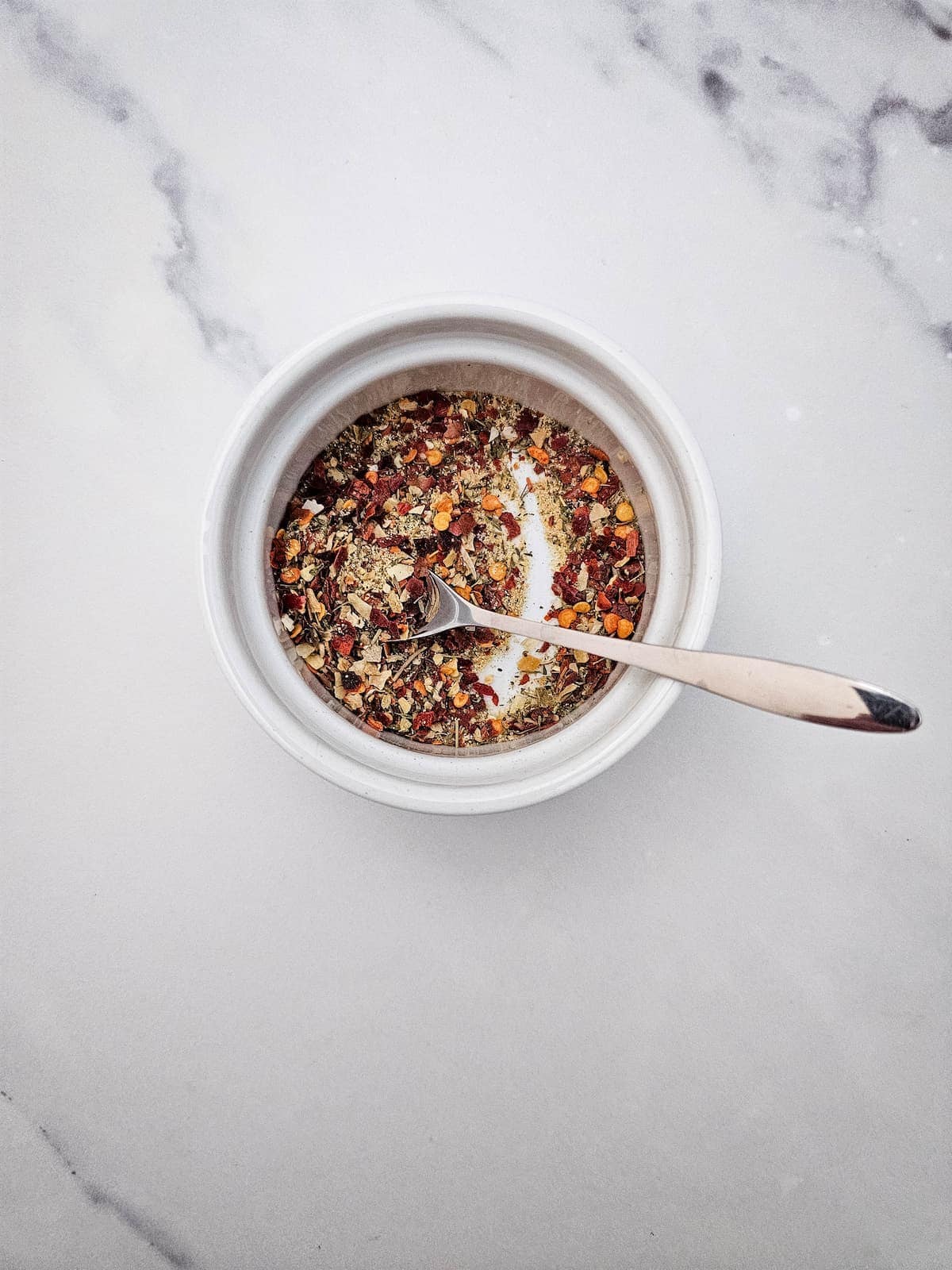 A small white bowl filled with mixed dried herbs and spices, with a metal spoon resting inside, on a marble surface.