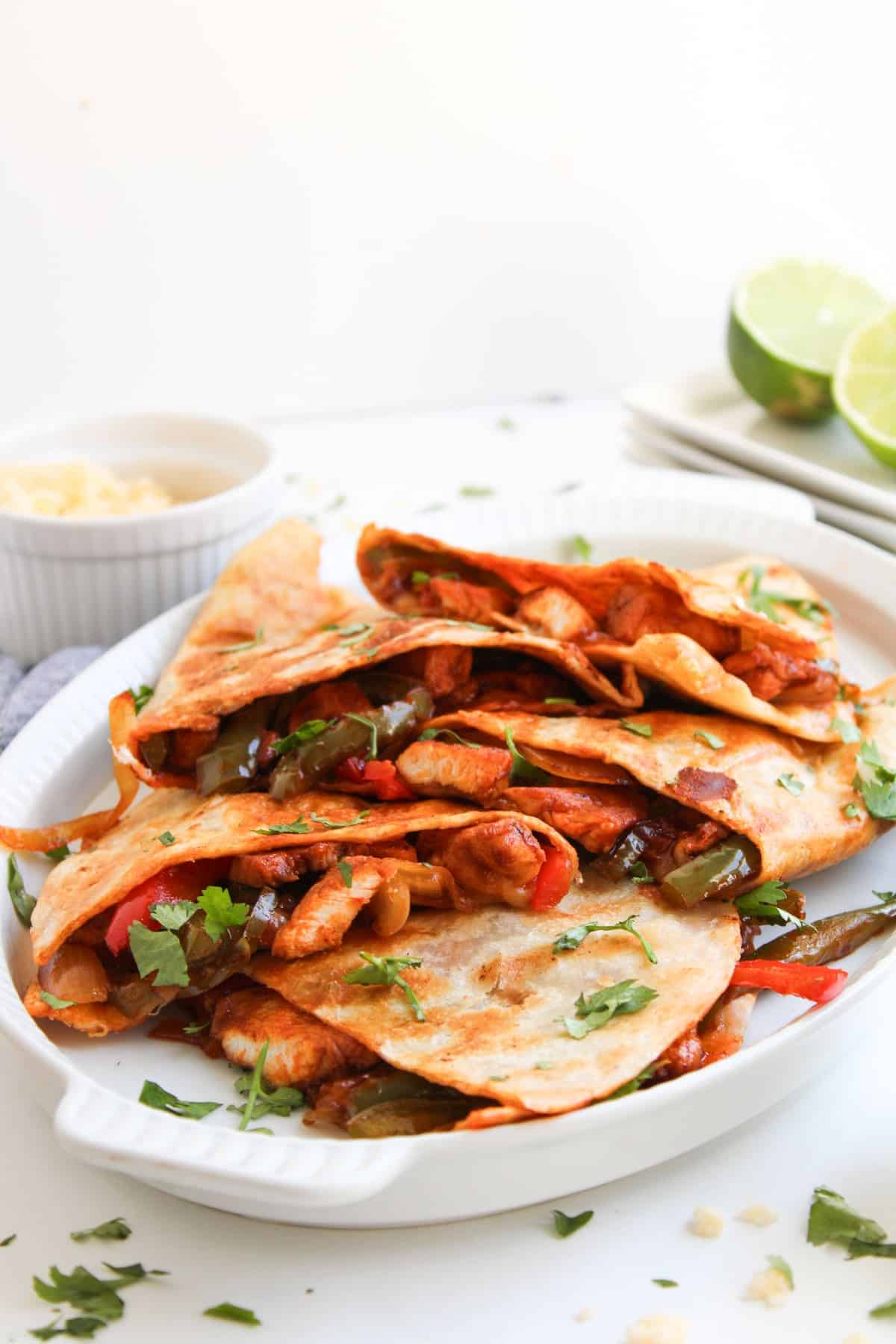 A white dish filled with folded chicken and vegetable quesadillas, garnished with chopped cilantro, with lime halves and a bowl of cheese in the background.