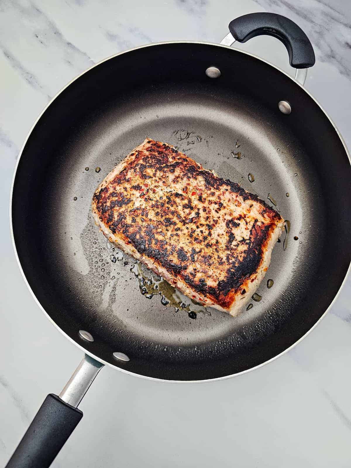 A piece of seared fish with a browned crust is cooking in a nonstick frying pan with some oil on a marble countertop.