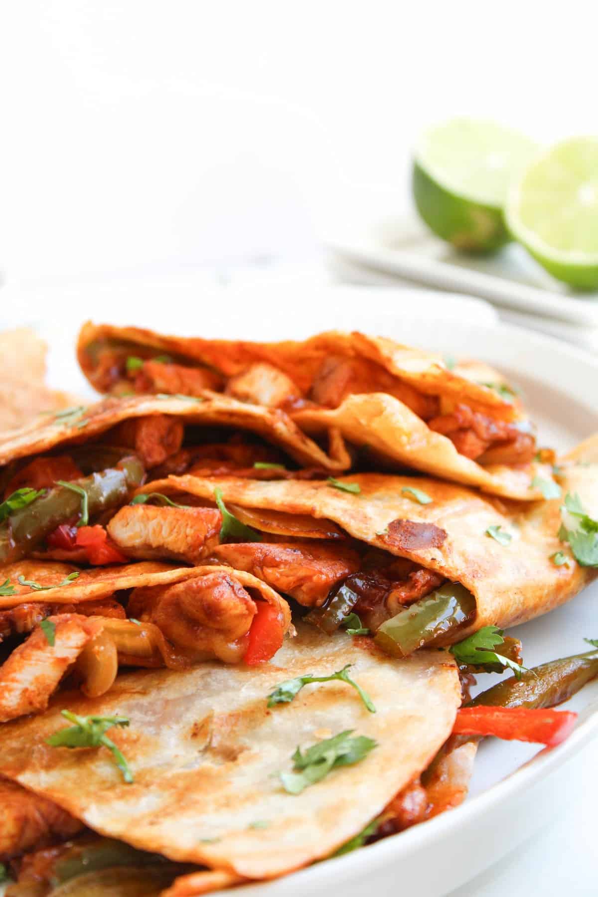 Three folded chicken quesadillas filled with sautéed peppers and onions are served on a plate, garnished with chopped cilantro. Lime halves are visible in the background.