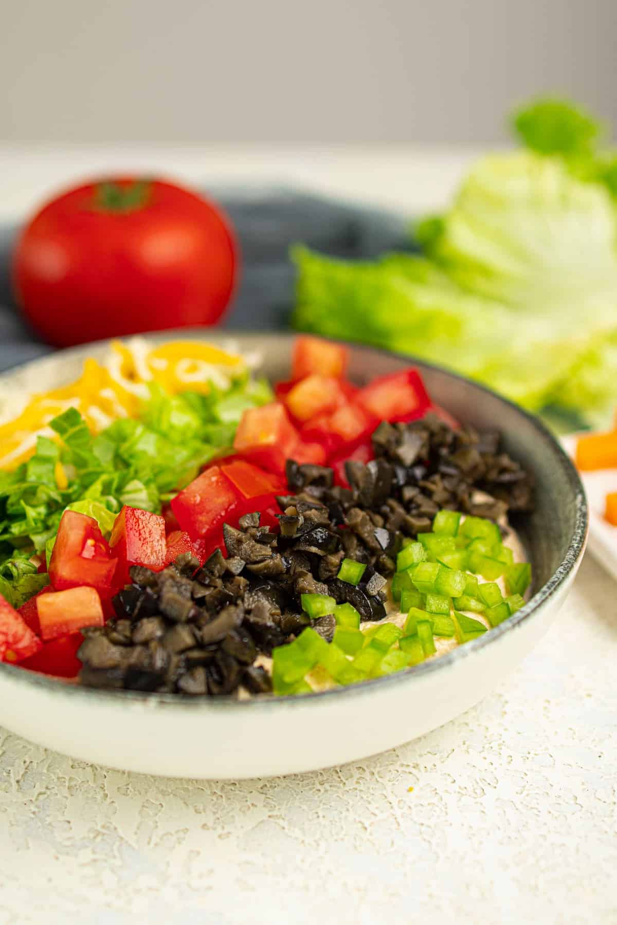 A bowl filled with chopped black olives, green bell peppers, tomatoes, green onions, and shredded cheese sits on a white surface, with a tomato and lettuce in the background.