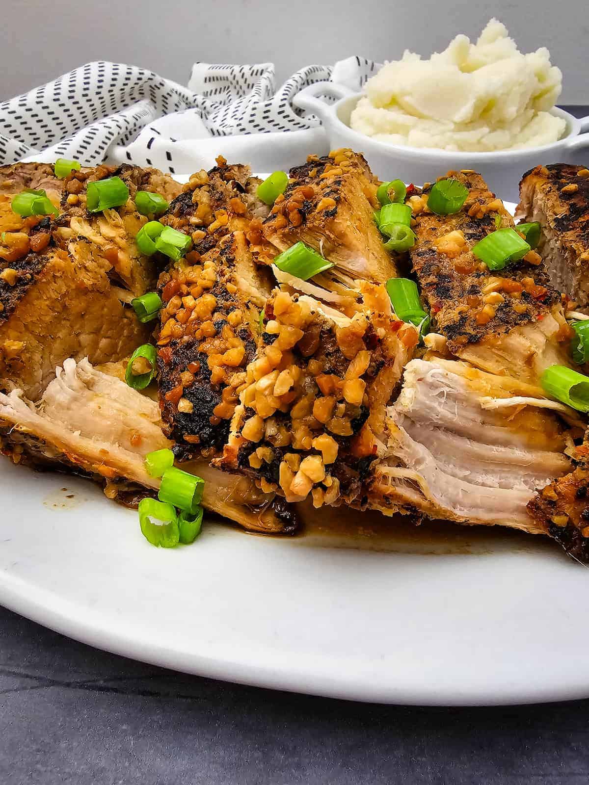 Sliced pork roast topped with chopped garlic and green onions on a white plate, with a bowl of mashed potatoes in the background.