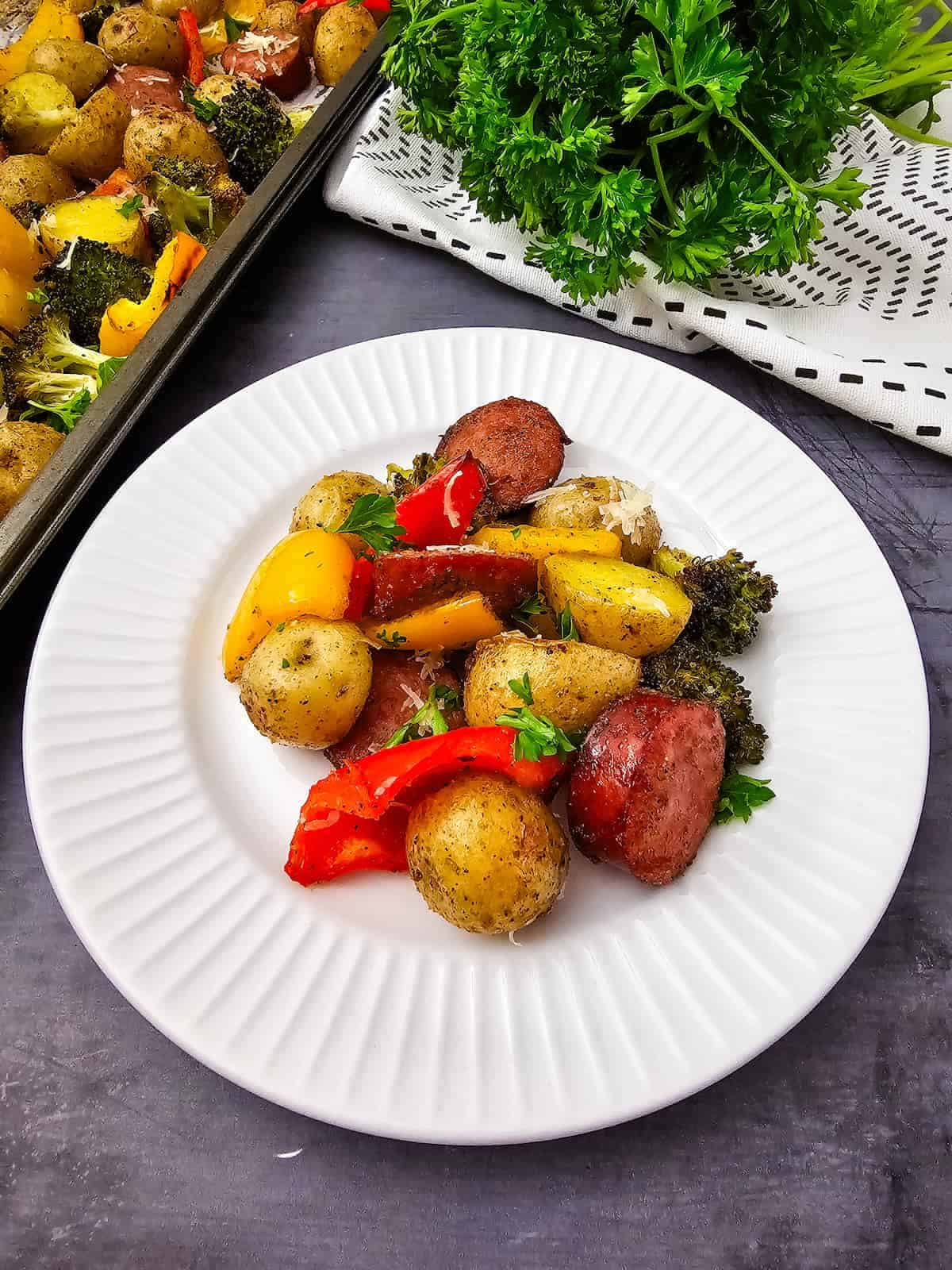 A white plate with roasted baby potatoes, sausage slices, broccoli, and assorted bell peppers, garnished with fresh parsley. A baking sheet and fresh herbs are in the background.