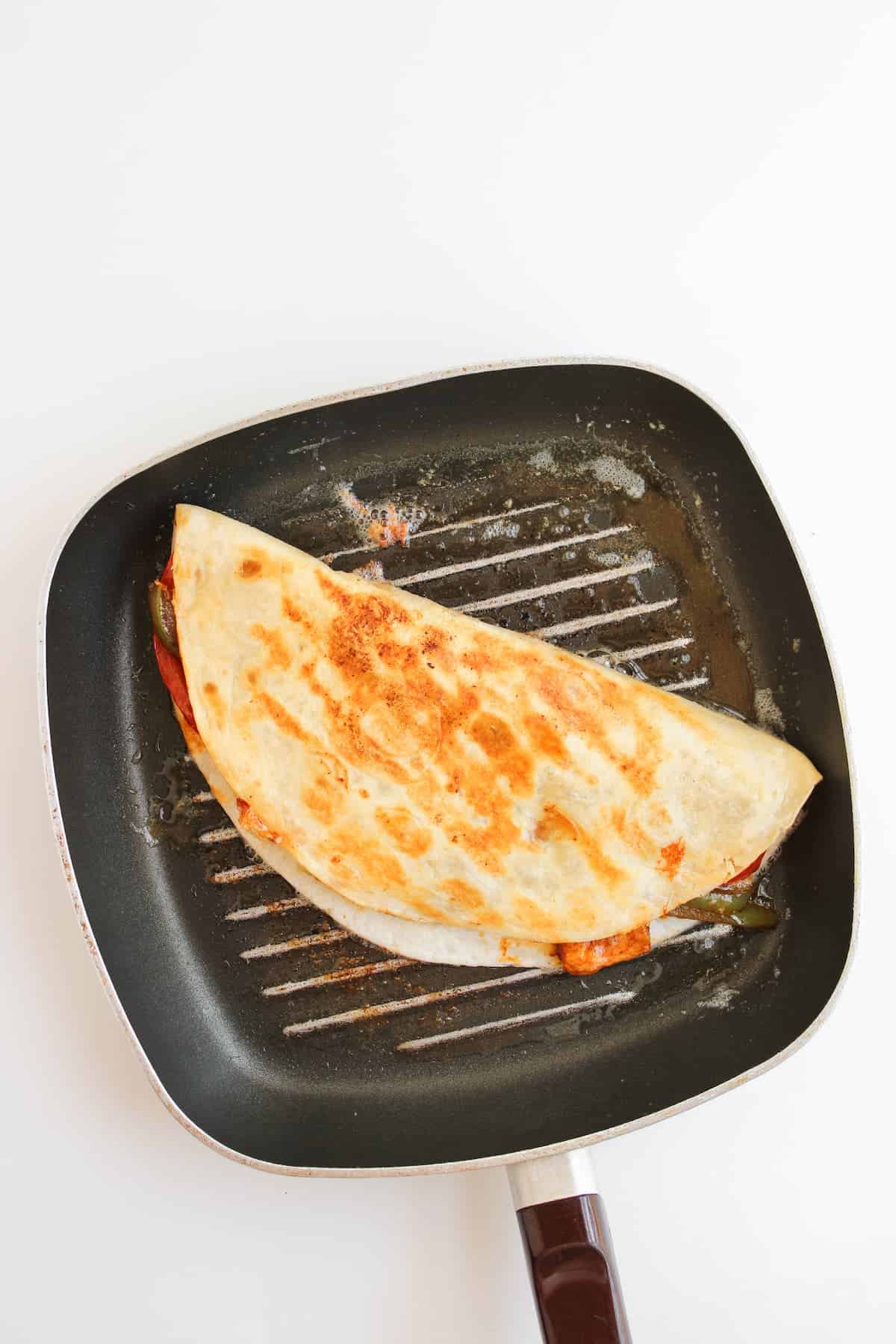 A folded quesadilla with a golden-brown crust is cooking in a square grill pan on a white background.