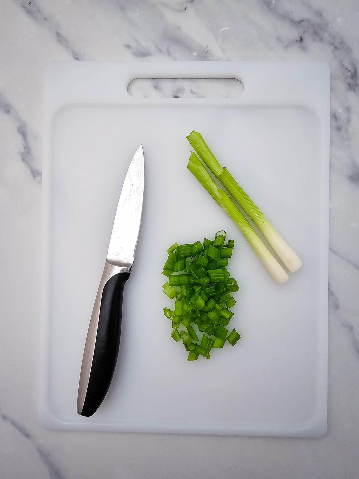 A paring knife, chopped green onions, and whole green onions on a white cutting board placed on a marble surface.