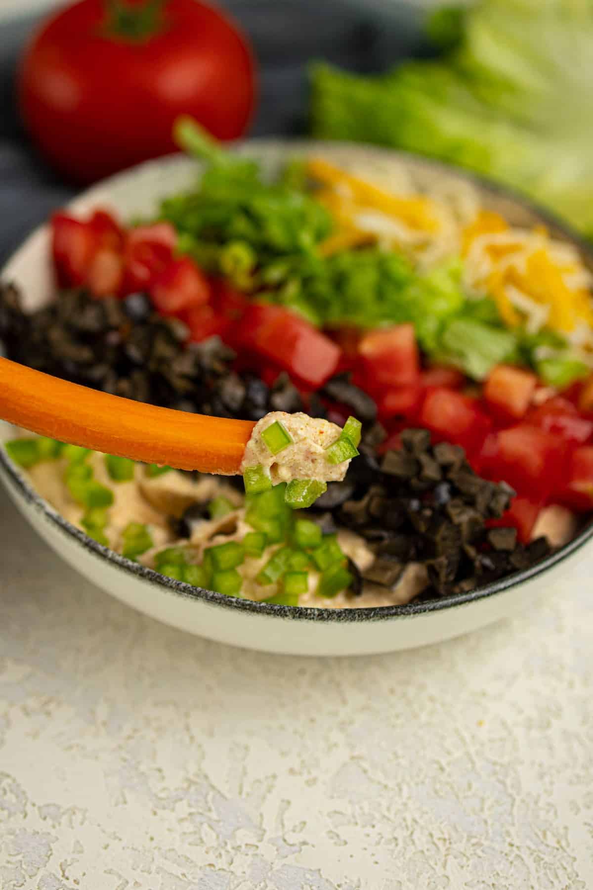 A carrot stick with dip and chopped vegetables held above a bowl of layered dip with diced tomatoes, olives, lettuce, and shredded cheese.