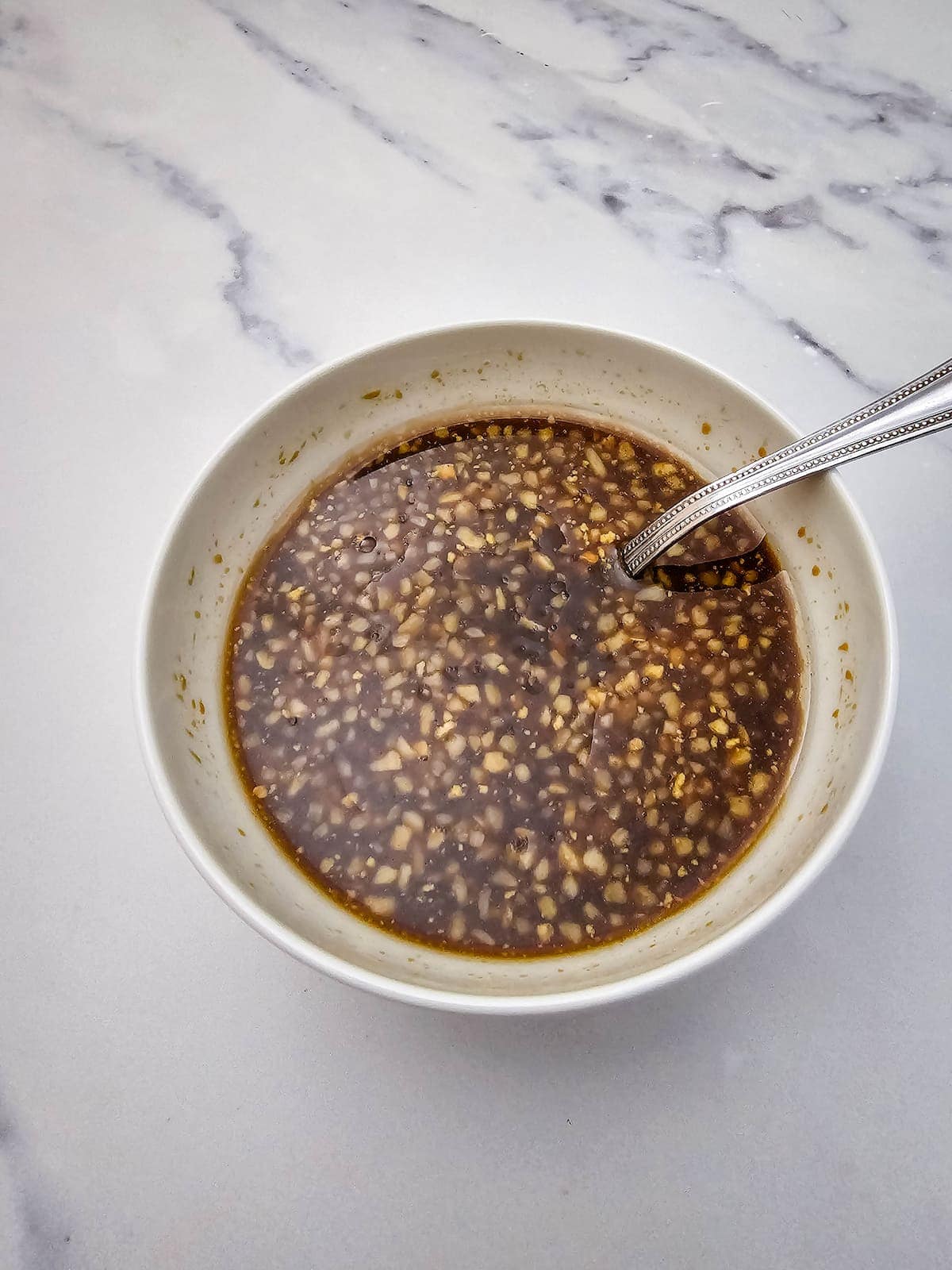 A white bowl with a silver spoon containing a chunky, brown liquid sauce with visible bits of garlic on a marble surface.
