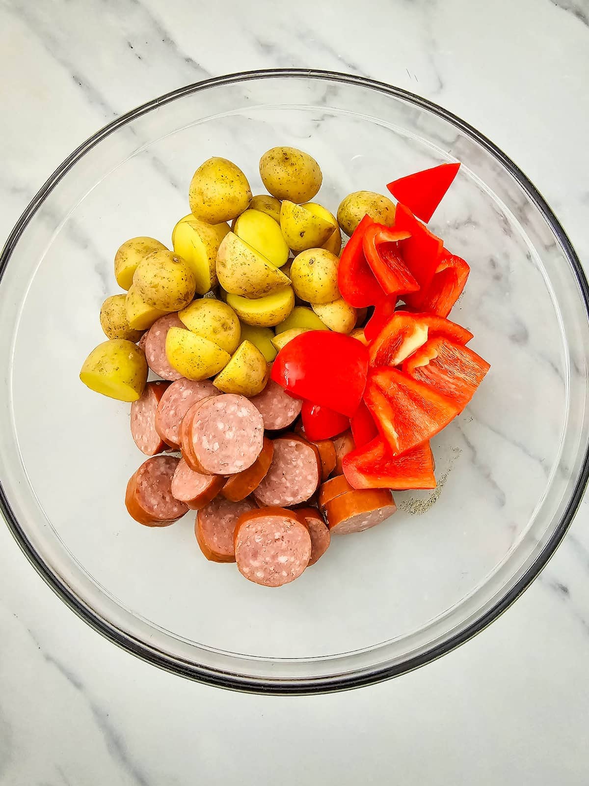 Clear glass bowl with sliced sausage, halved baby potatoes, and chopped red bell pepper on a white marble surface.