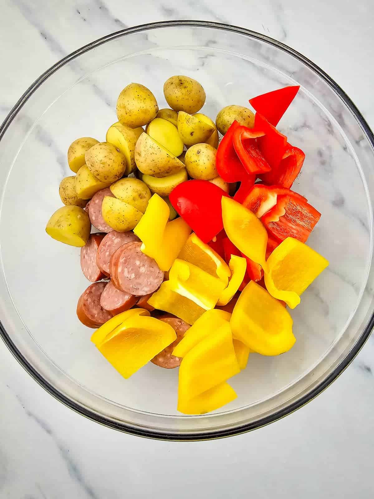 A glass bowl contains sliced baby potatoes, yellow bell peppers, red bell peppers, and sausage pieces on a white marble surface.