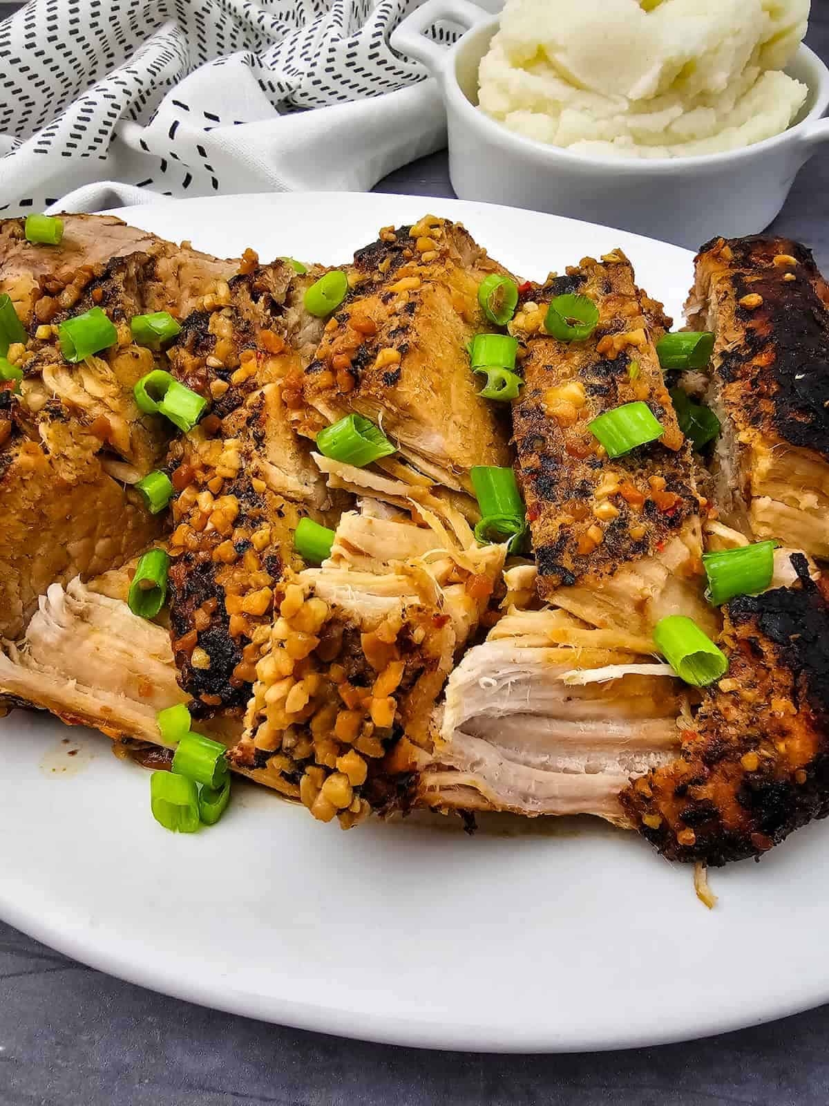 Sliced, seasoned pork ribs garnished with chopped green onions on a white plate, with a bowl of mashed potatoes in the background.