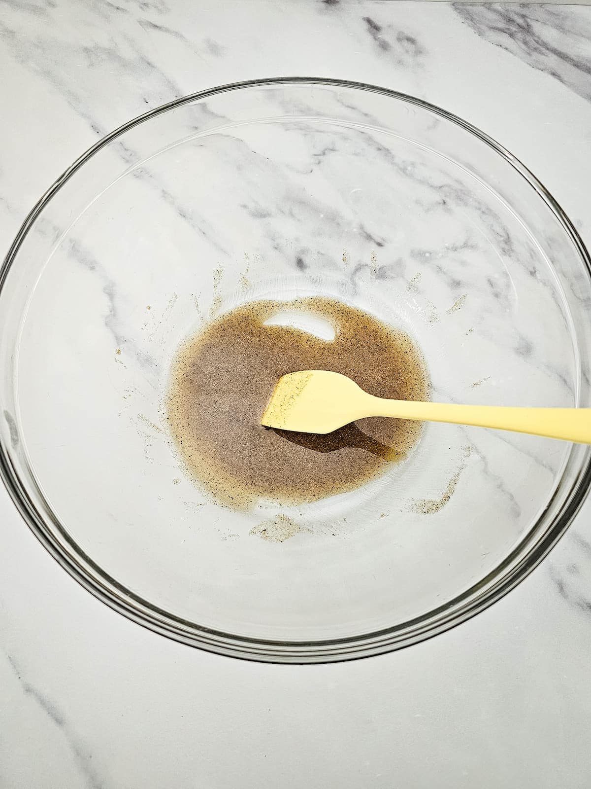 A glass mixing bowl containing a small amount of brown liquid being stirred with a yellow spatula on a white marble surface.