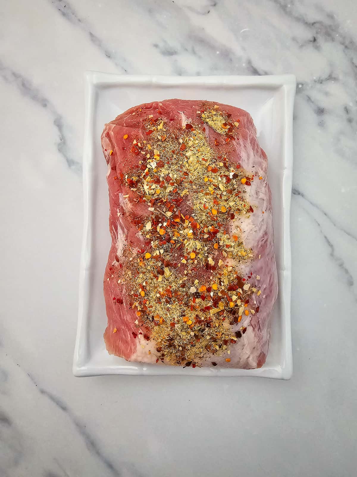 A raw pork loin topped with mixed spices and seasonings sits on a rectangular white plate atop a marble surface.